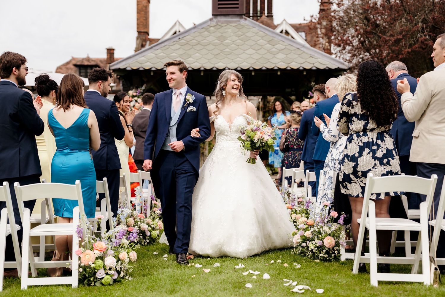 Newlyweds walking back down aisle surrounded by applauding guests and floral petal decorations.