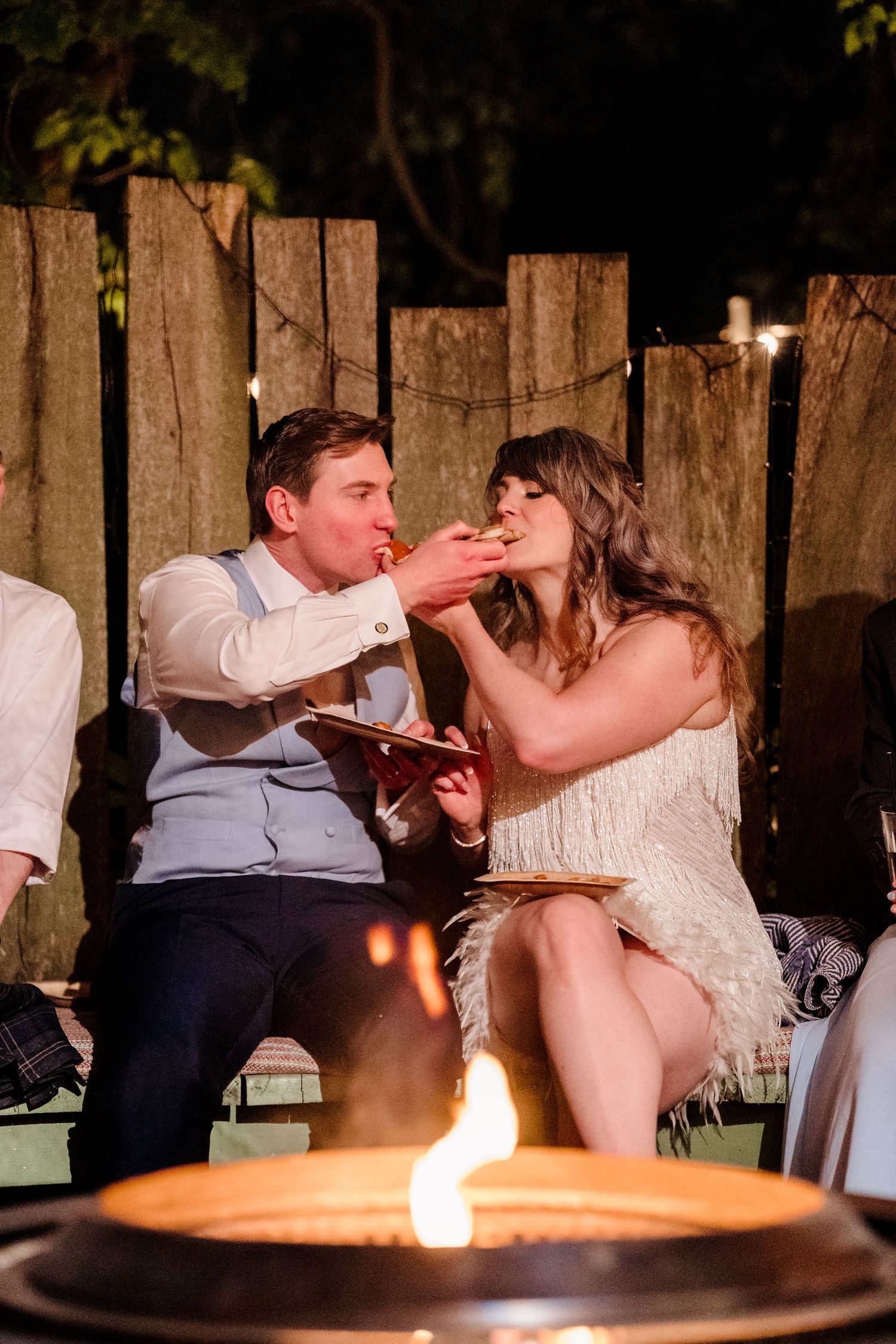 Couple feeding each other snacks by a fire pit at an outdoor evening wedding celebration.