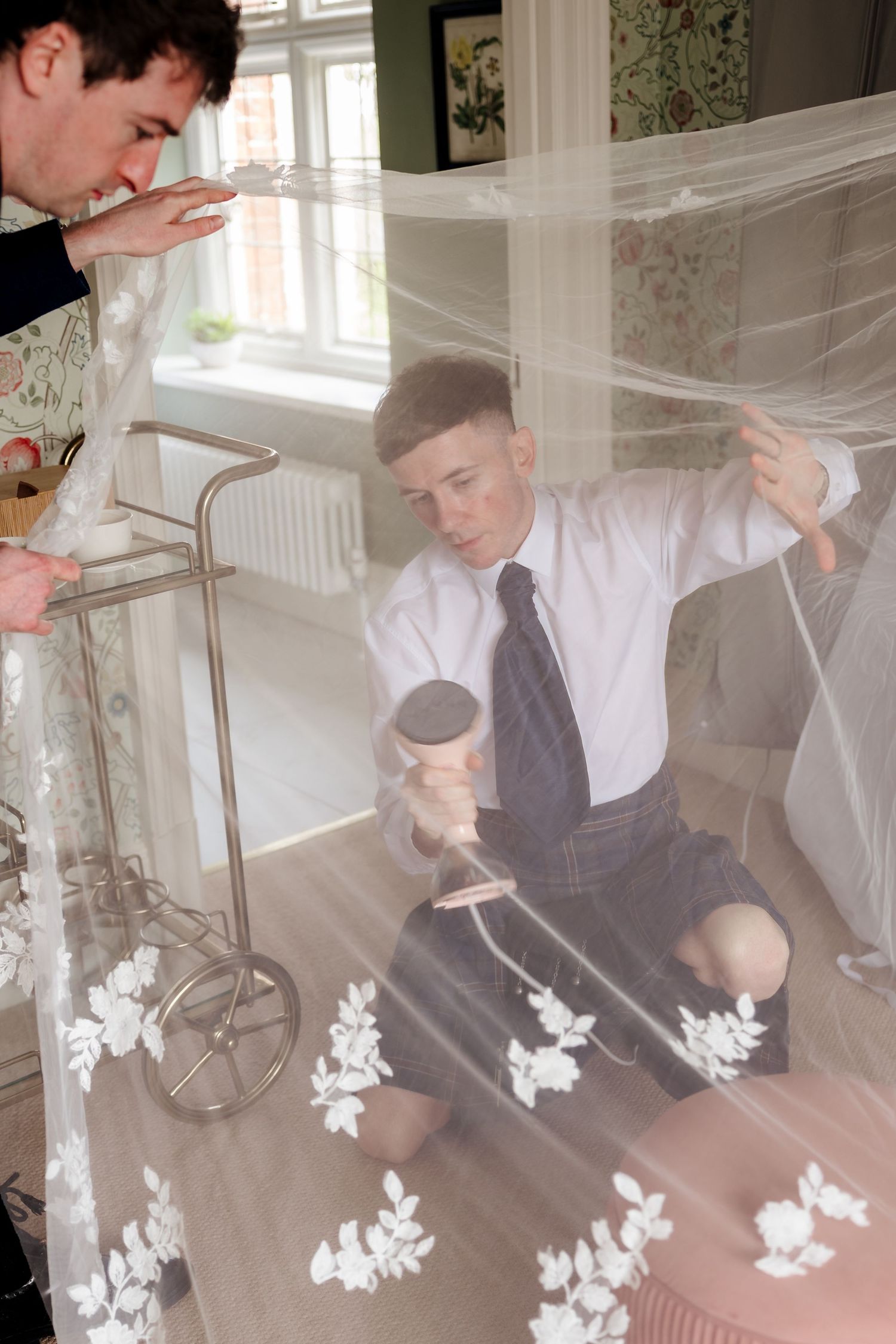 Two people steaming a floral wedding veil indoors, with one kneeling holding a steamer beneath the delicate fabric.