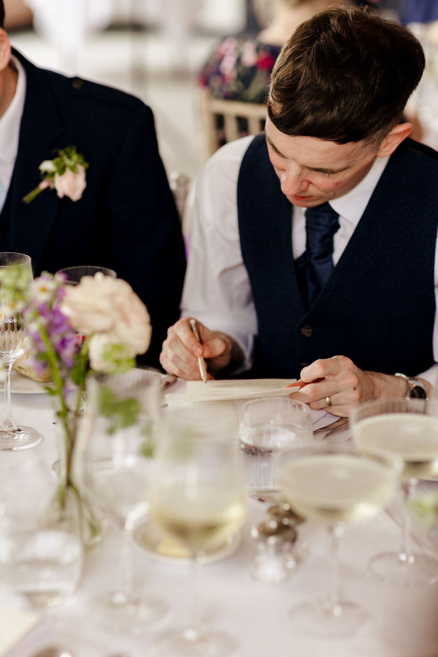 Groom in navy vest signing wedding documents at a reception table with floral centerpieces and wine glasses.