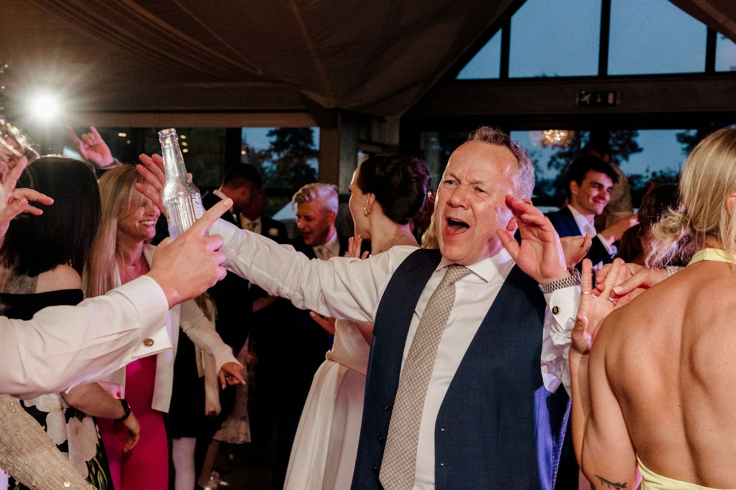 Joyful bald man dancing and singing at a lively wedding reception surrounded by guests on the dance floor.