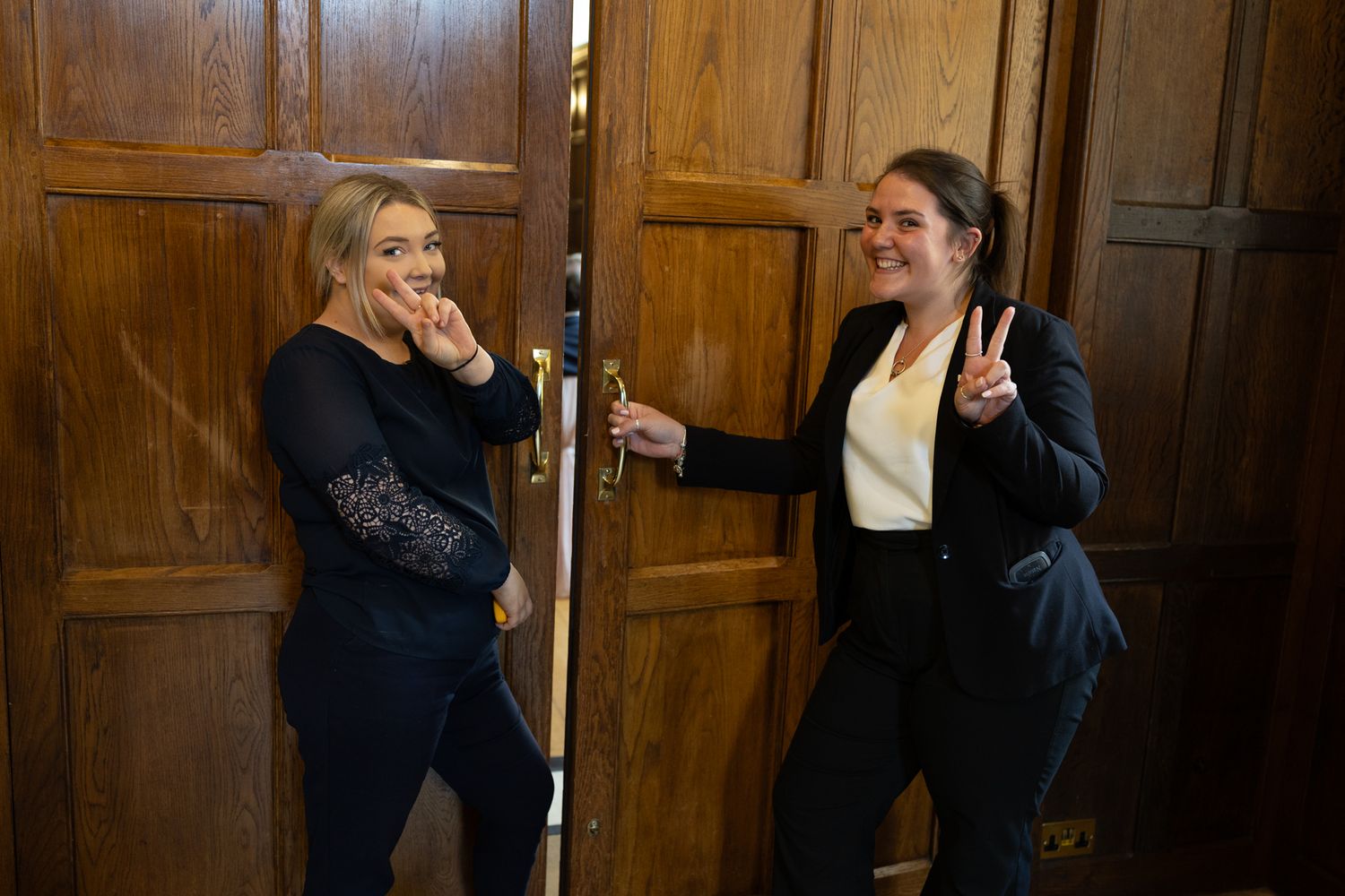 Two women posing by ornate wooden doors, one making a kiss face and the other giving a peace sign.