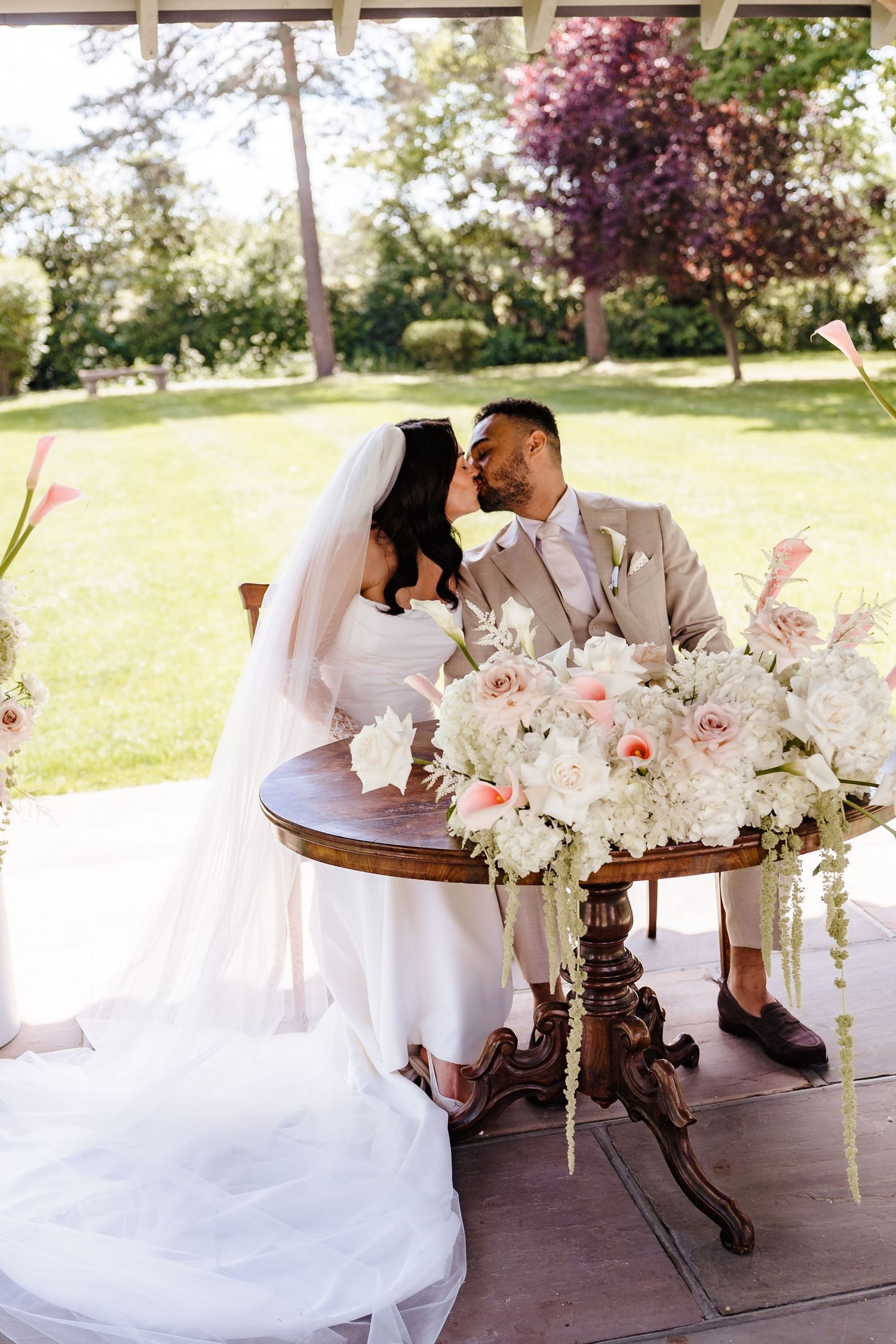 Bride and groom share a kiss at signing table adorned with white and blush floral arrangements during outdoor wedding ceremony.