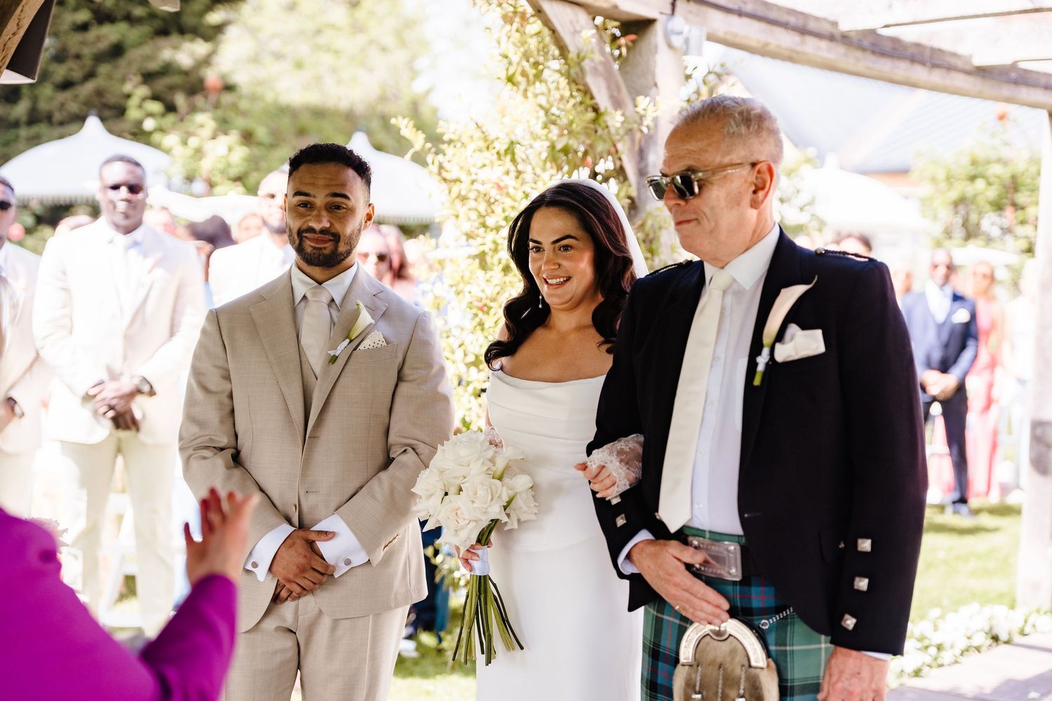 Bride in white gown walks down aisle with groom in tan suit and older man in Scottish kilt at outdoor wedding.