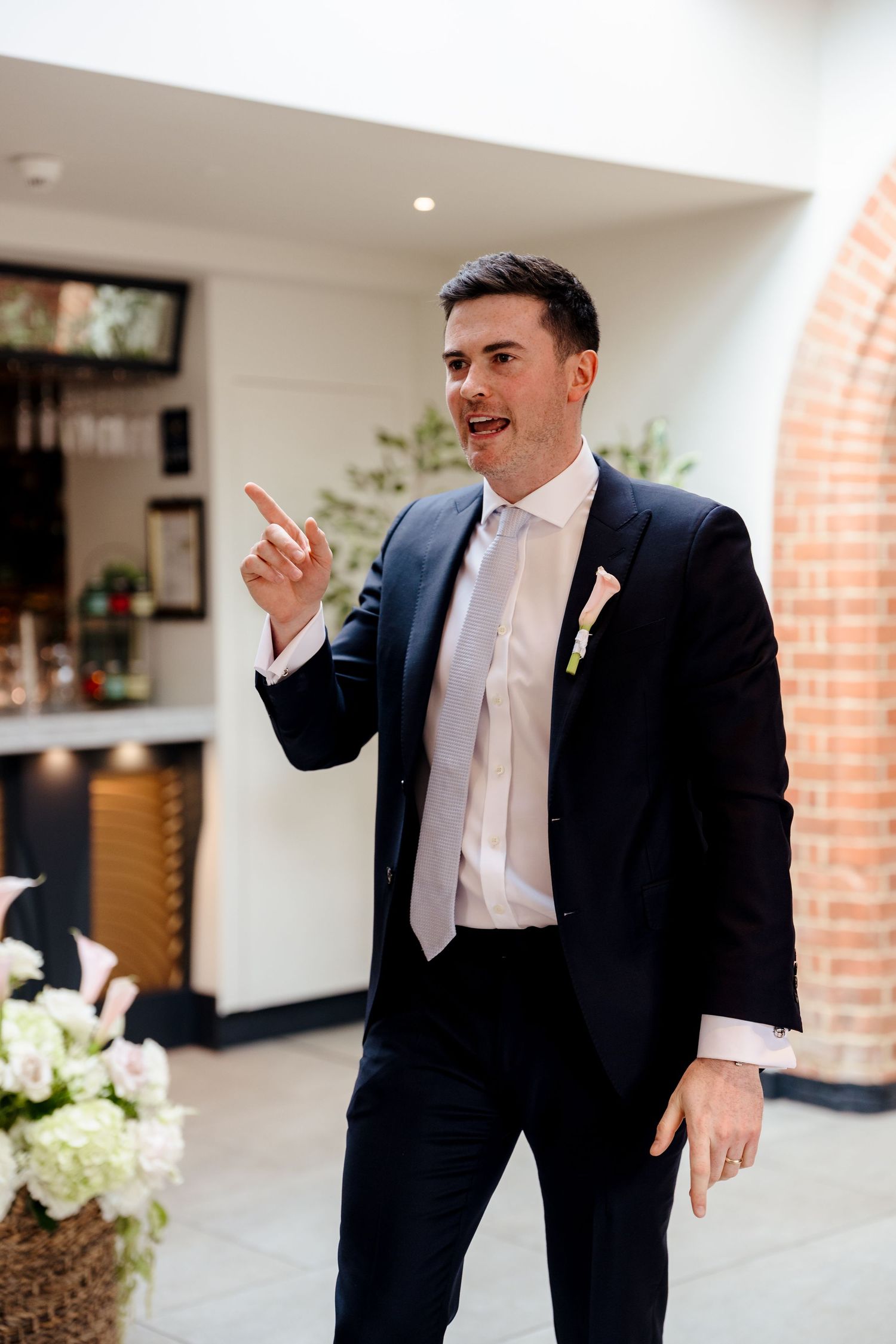 Dapper man in navy suit with pink boutonniere gestures and smiles during indoor wedding reception speech.