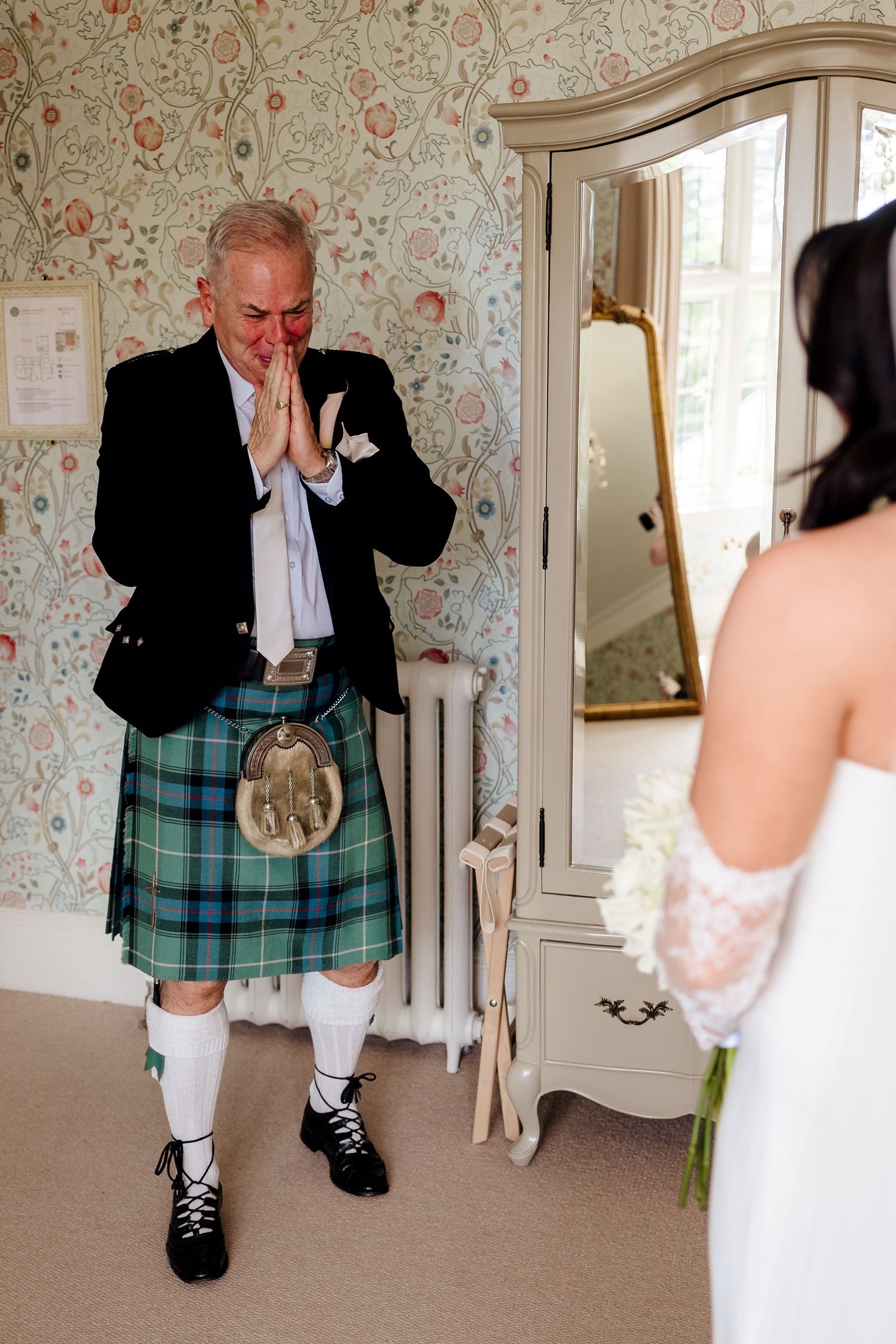 Emotional father in Scottish kilt clasps hands in awe seeing bride daughter on wedding day indoors.