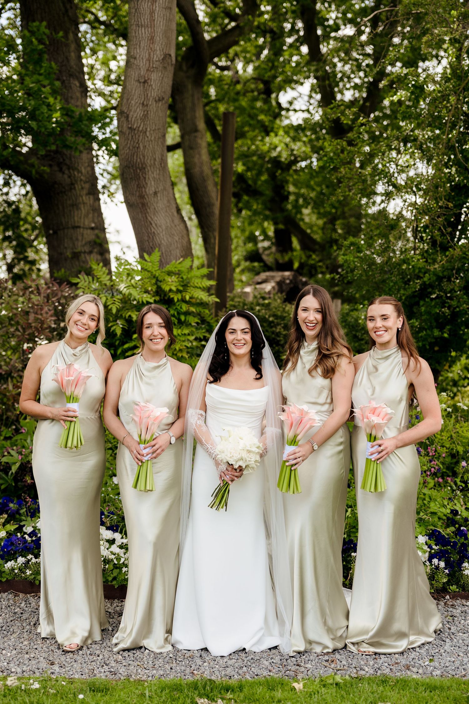 Bride in white gown with four bridesmaids in sage satin dresses holding pink bouquets in a garden.