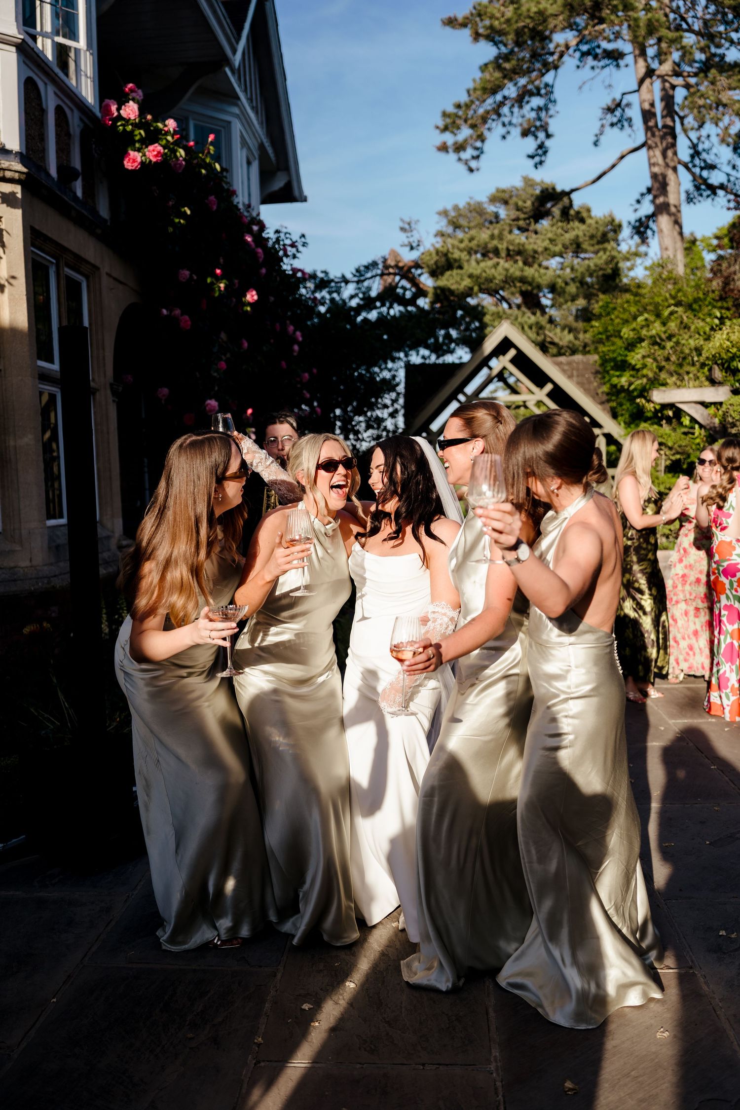 Bride in white dress laughs joyfully with bridesmaids in sage green satin gowns outdoors.