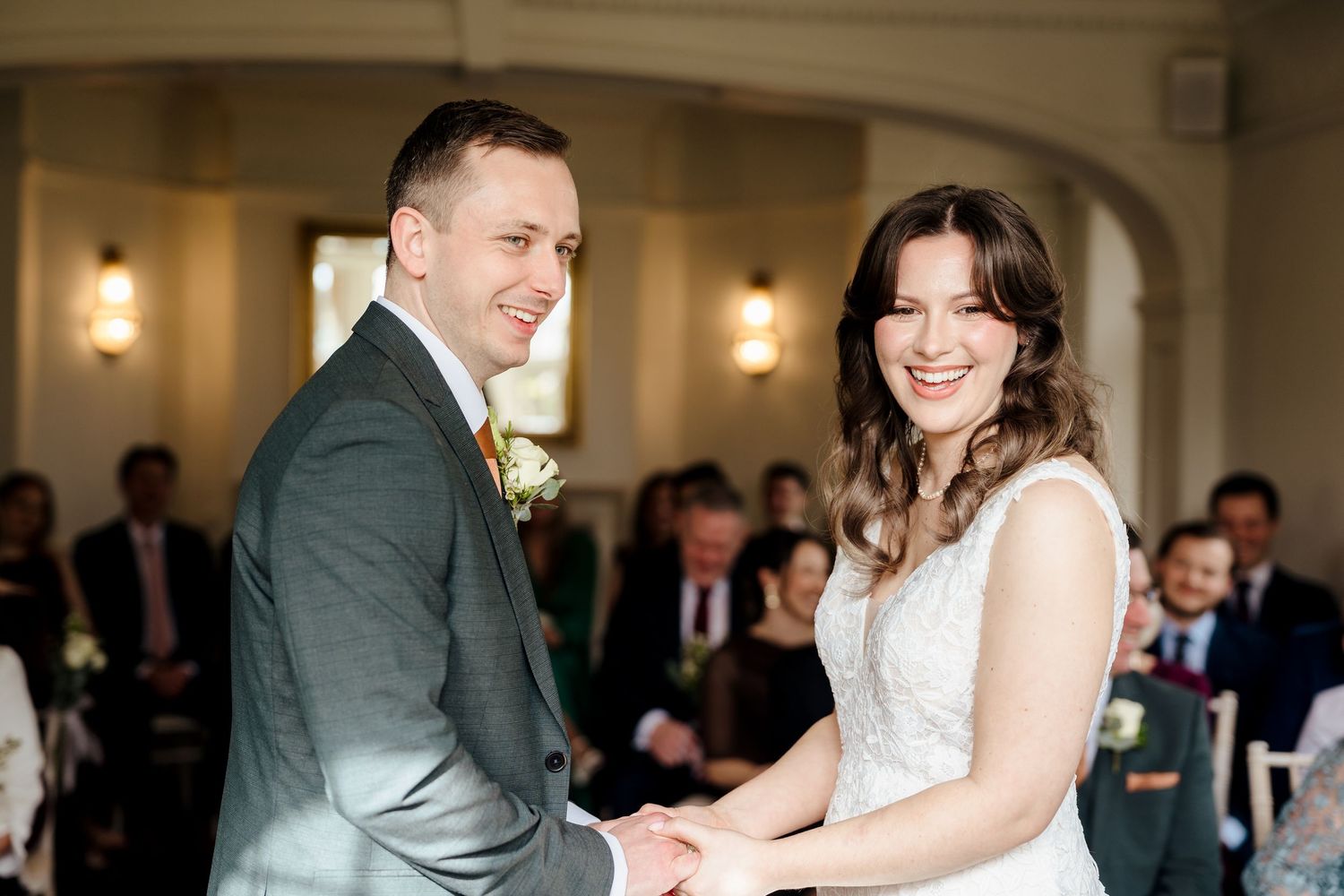 Bride and groom smiling joyfully while holding hands during their wedding ceremony in an elegant venue.