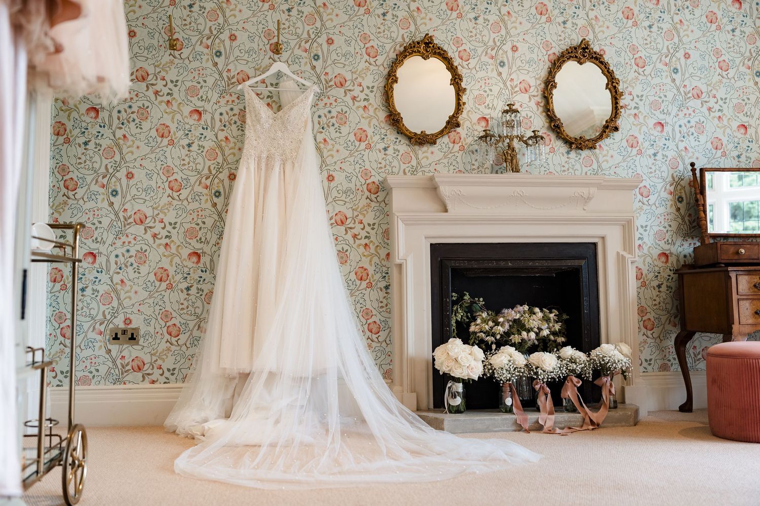 Elegant wedding dress hanging above a fireplace in a floral wallpapered room with bouquets and gold mirrors.