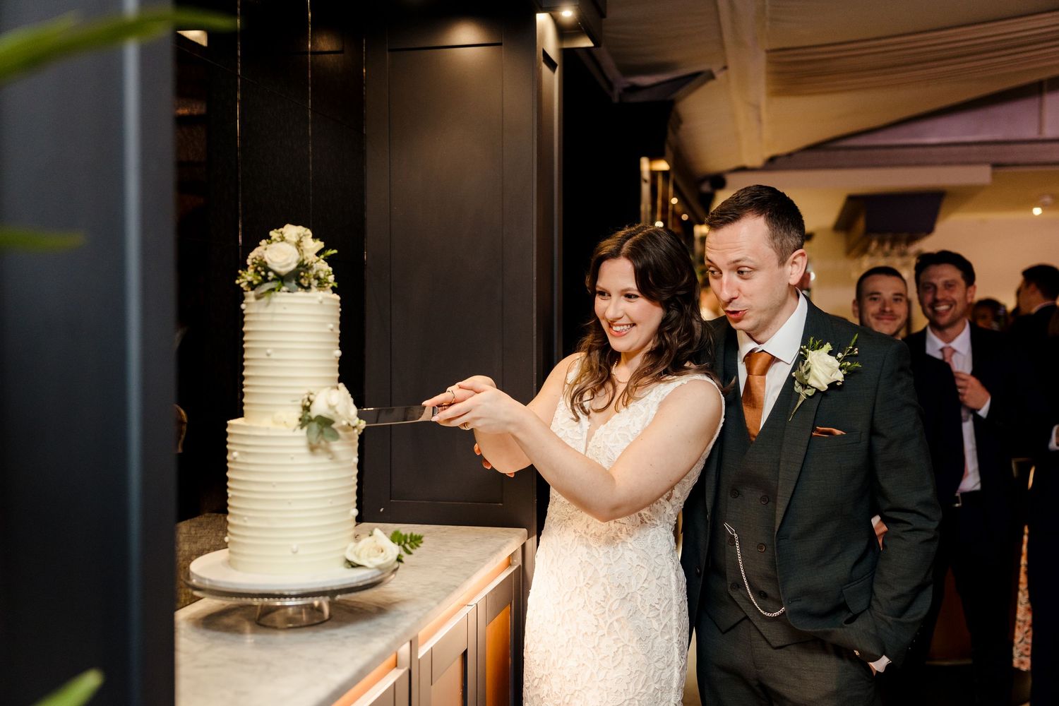 Smiling bride and groom cut elegant white tiered wedding cake at indoor reception.