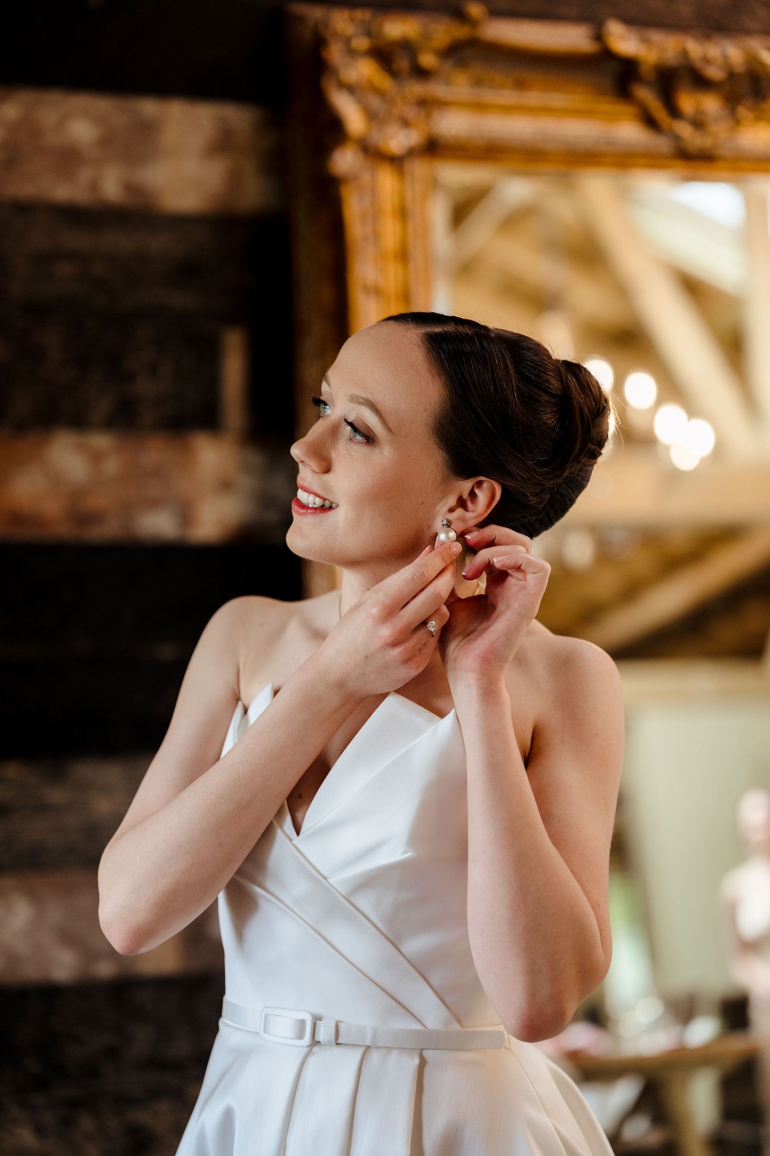 Bride smiling while putting on earrings in a rustic barn venue wearing a white halter wedding dress.