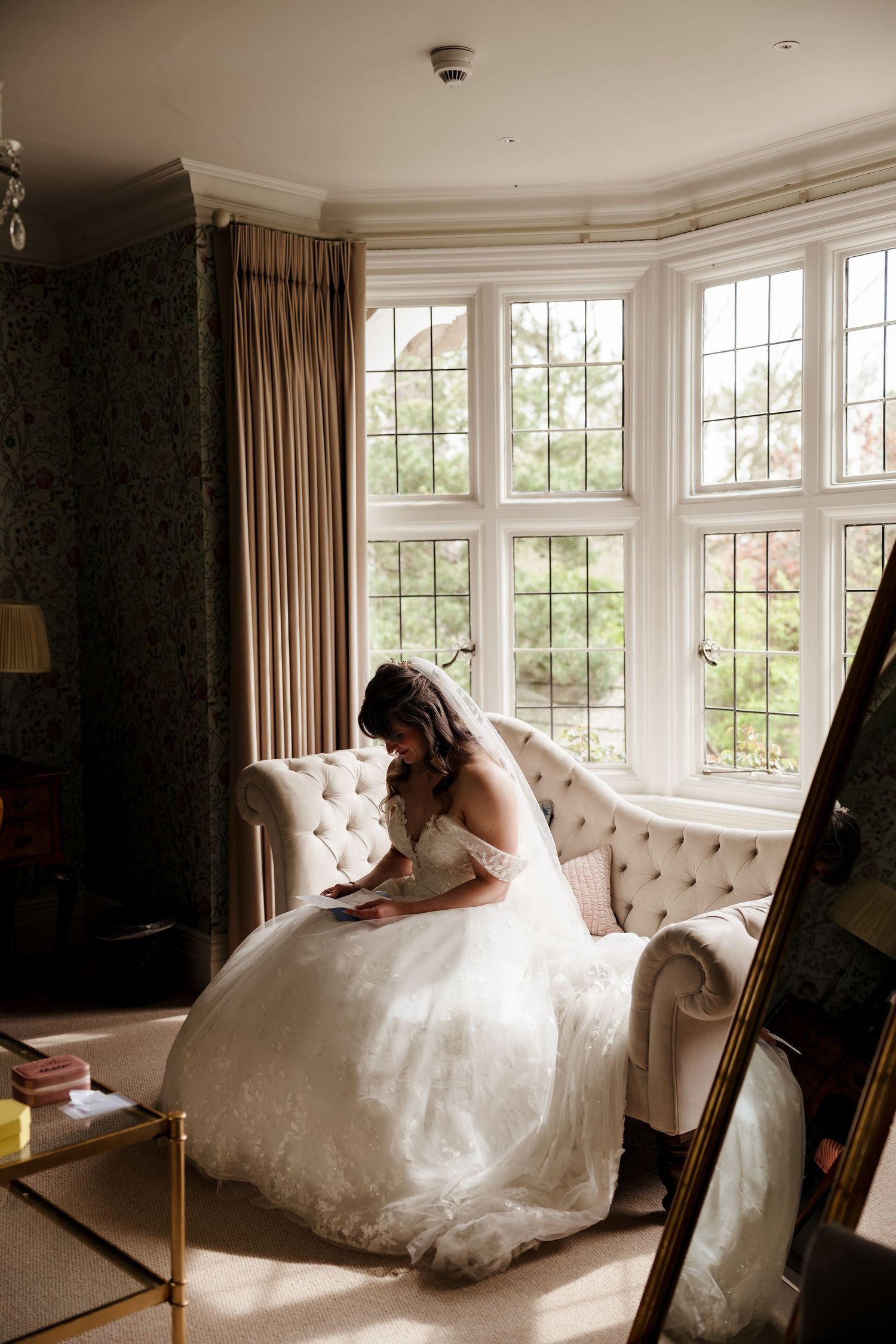 Bride in white wedding gown sitting on tufted sofa in elegant bay window room.
