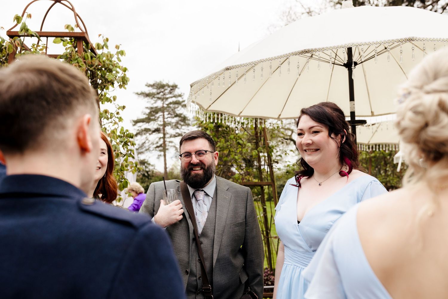 Bride in light blue dress laughing with bearded officiant during outdoor wedding ceremony under white umbrella.