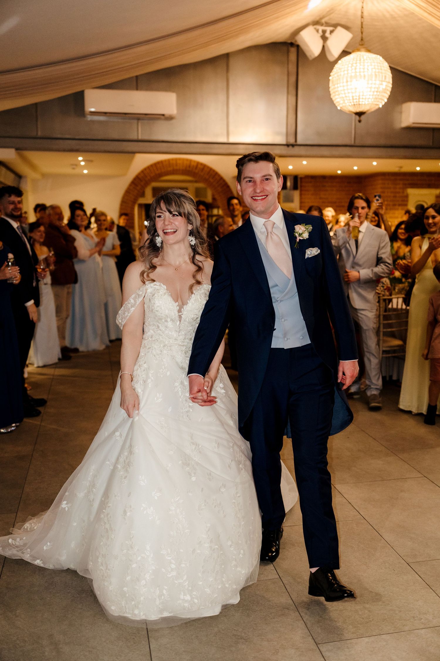 Bride and groom walk hand-in-hand into their wedding reception, smiling joyfully as guests celebrate around them.