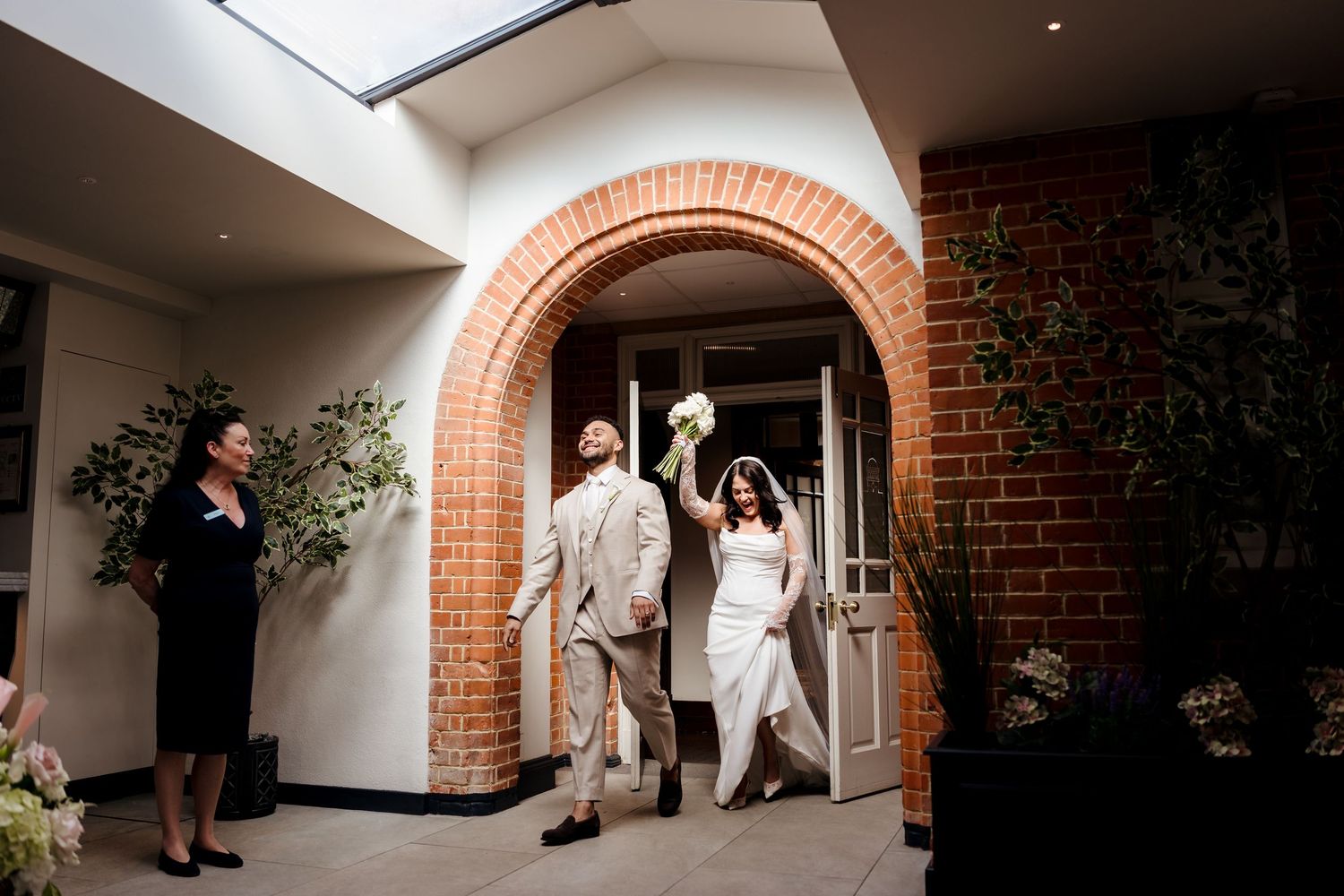 Bride and groom entering wedding venue through brick archway, bouquet raised.