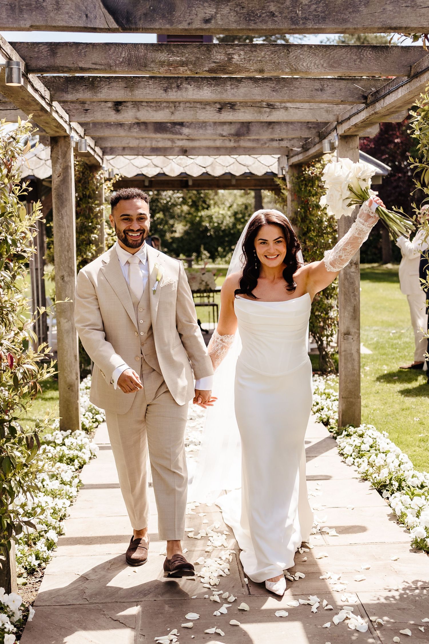Joyful newlyweds walking hand in hand under pergola arch, bride raising bouquet in celebration outdoors.