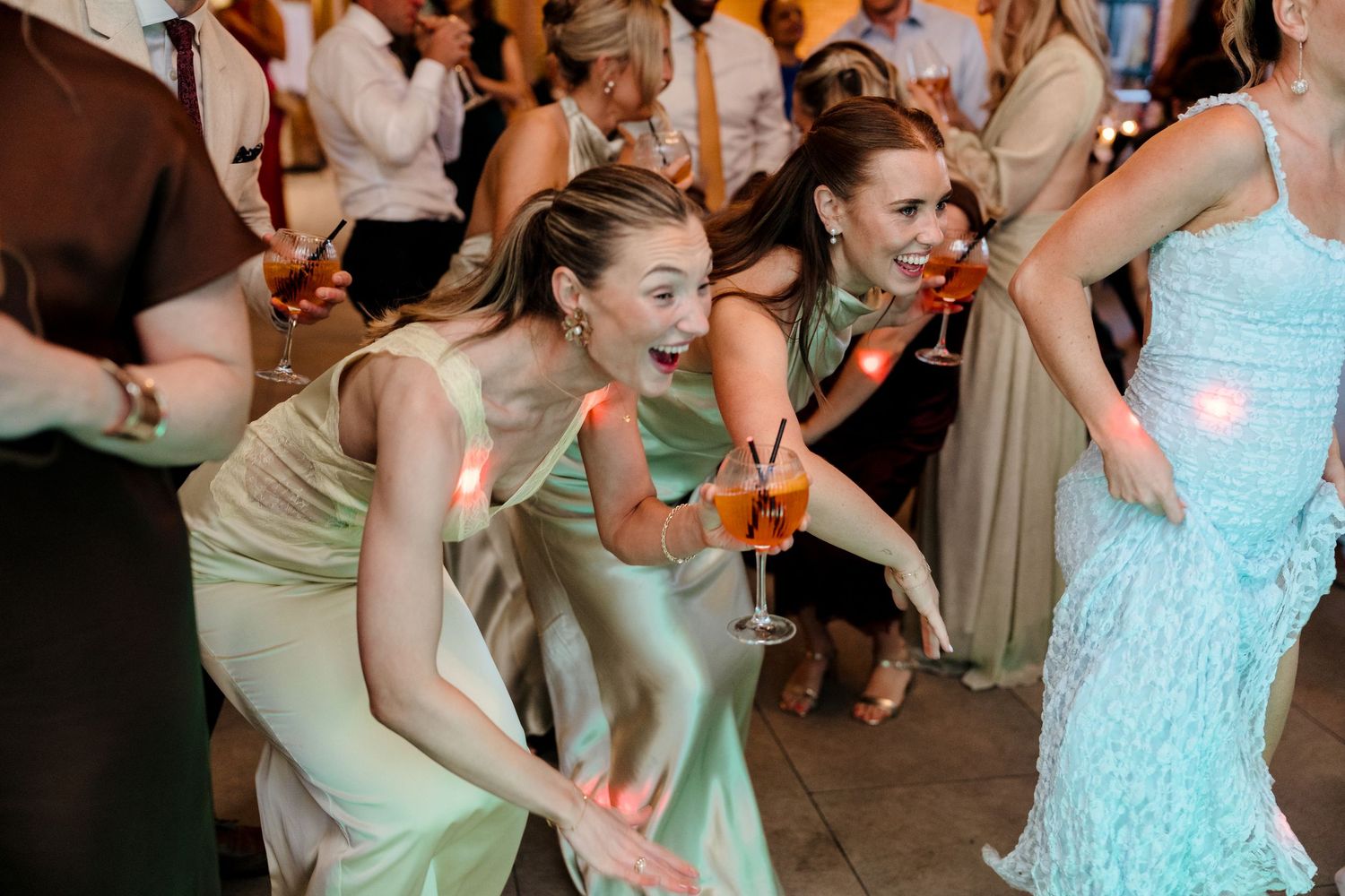 Bridesmaids in sage green satin dresses dancing energetically with cocktails at lively wedding reception.