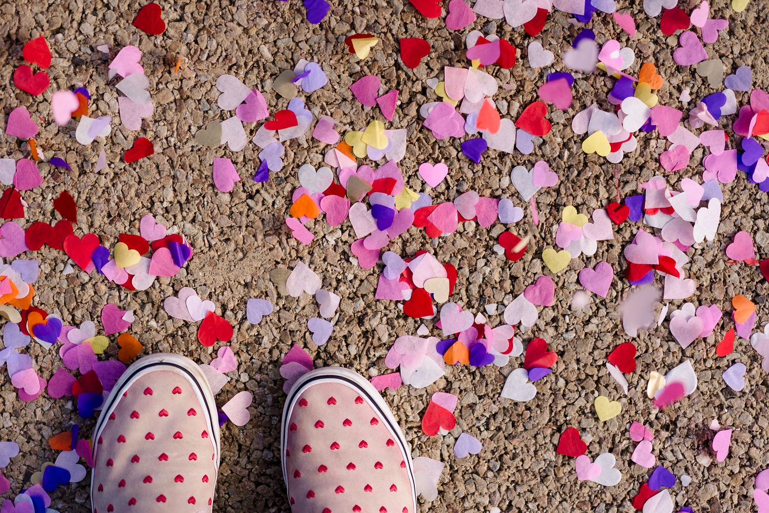 Pink sneakers with heart prints standing on gravel surrounded by colorful heart-shaped confetti.