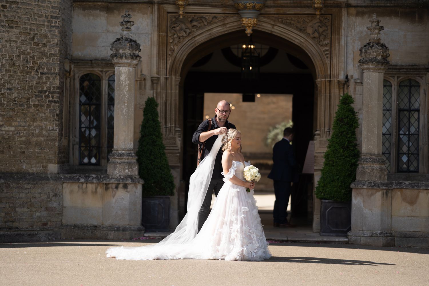 Bride and groom posing outside a grand stone building entrance, bride in white lace gown holding bouquet.