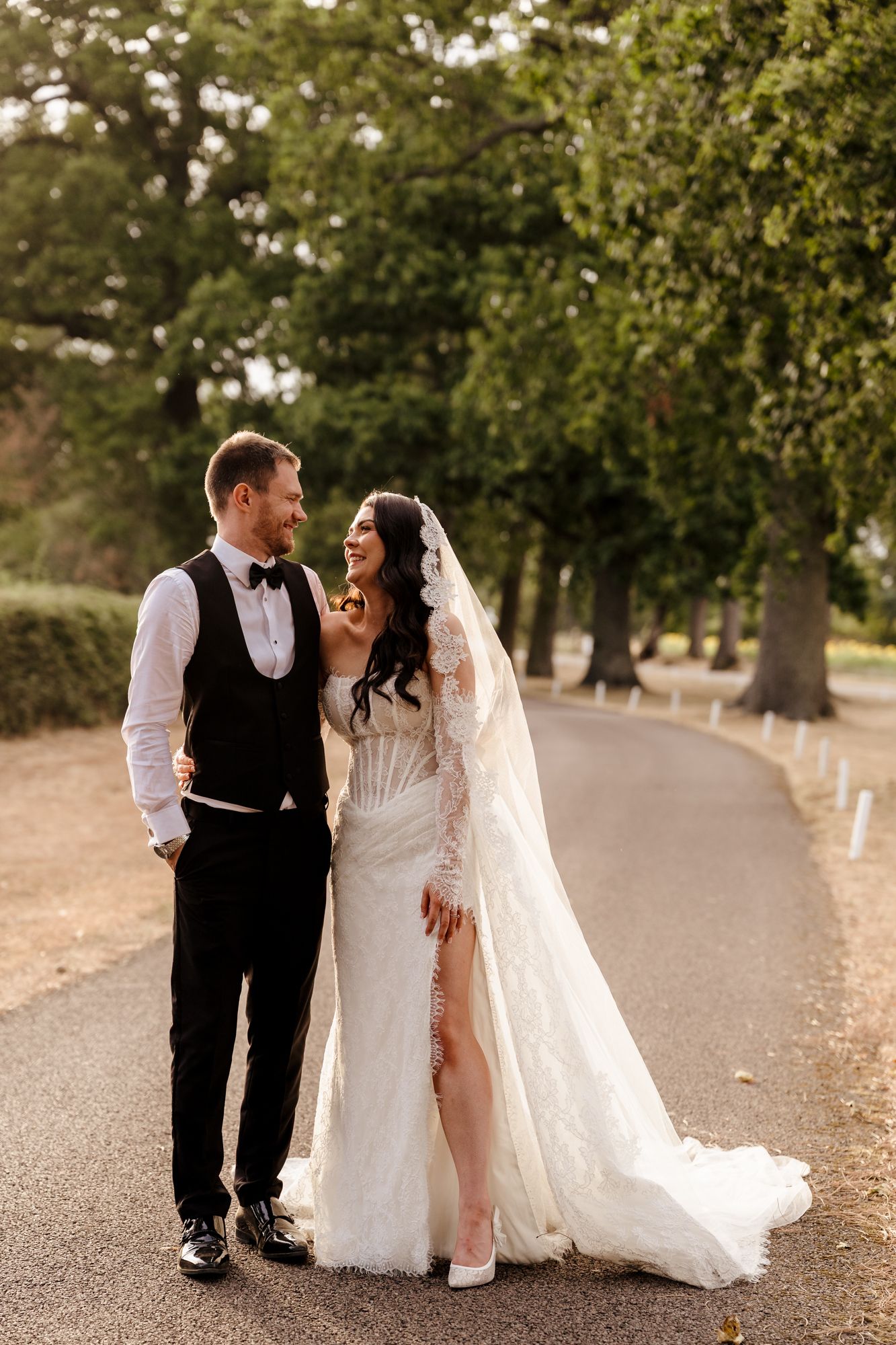 Bride and groom walking hand in hand down a tree-lined path, bride in lace gown with veil, groom in black tuxedo.