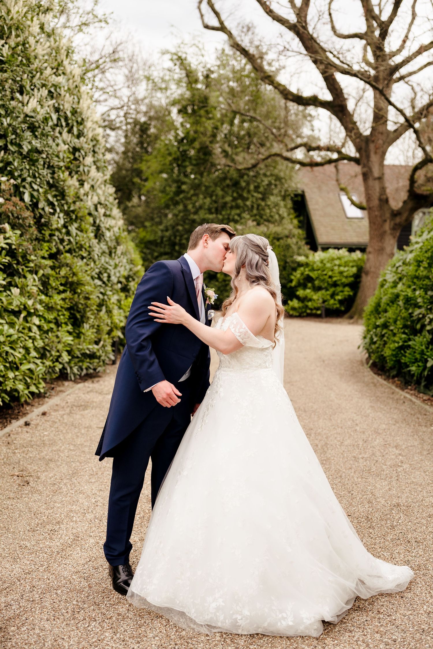 Bride and groom share a romantic kiss on a gravel garden path surrounded by lush greenery.