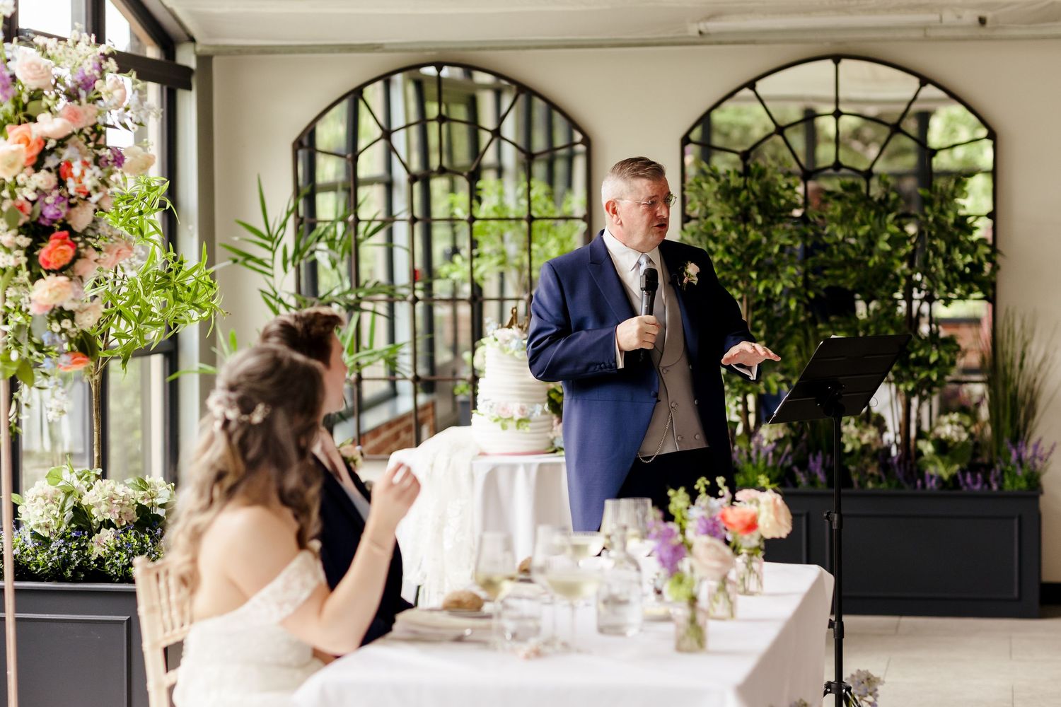 Father of the bride giving a speech at the wedding reception while the bride smiles at the table.