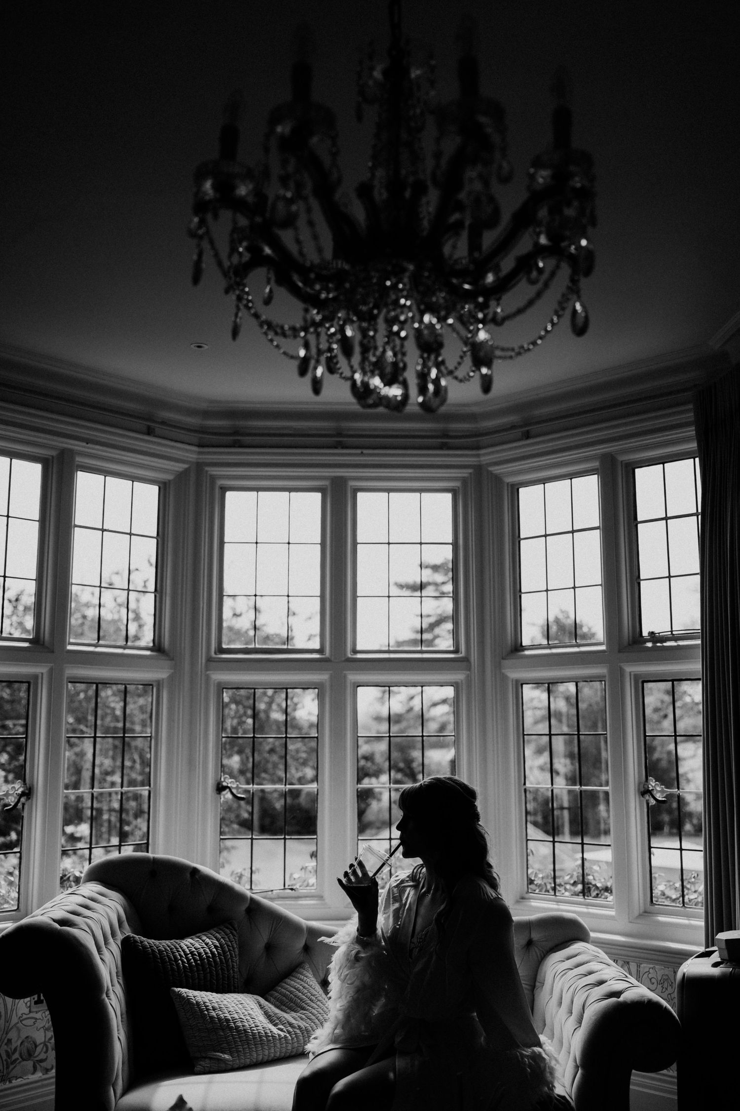 Black and white silhouette of a bride sitting by bay windows under a crystal chandelier in an elegant room.
