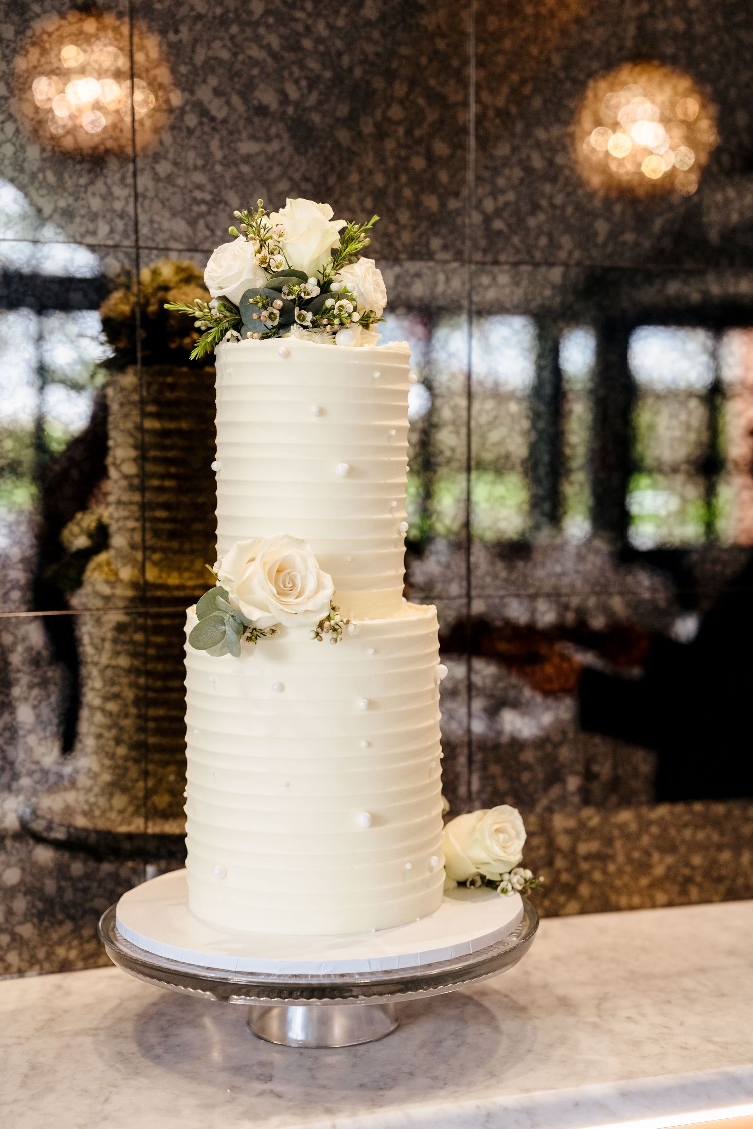 Elegant two-tier white wedding cake with ribbed frosting and white rose floral decorations on a silver stand.
