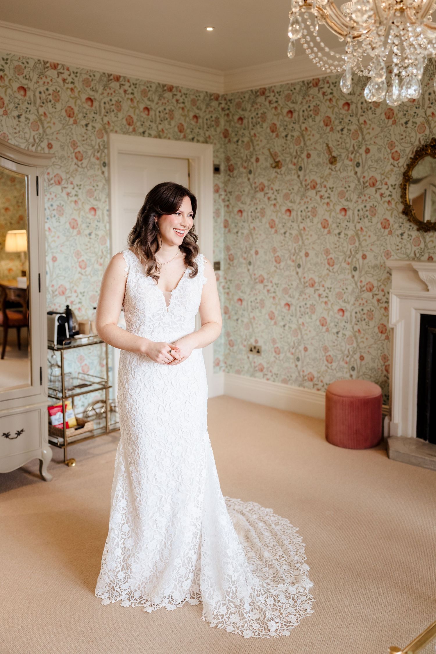 Bride in white lace wedding gown smiling in elegant floral wallpaper room with chandelier.
