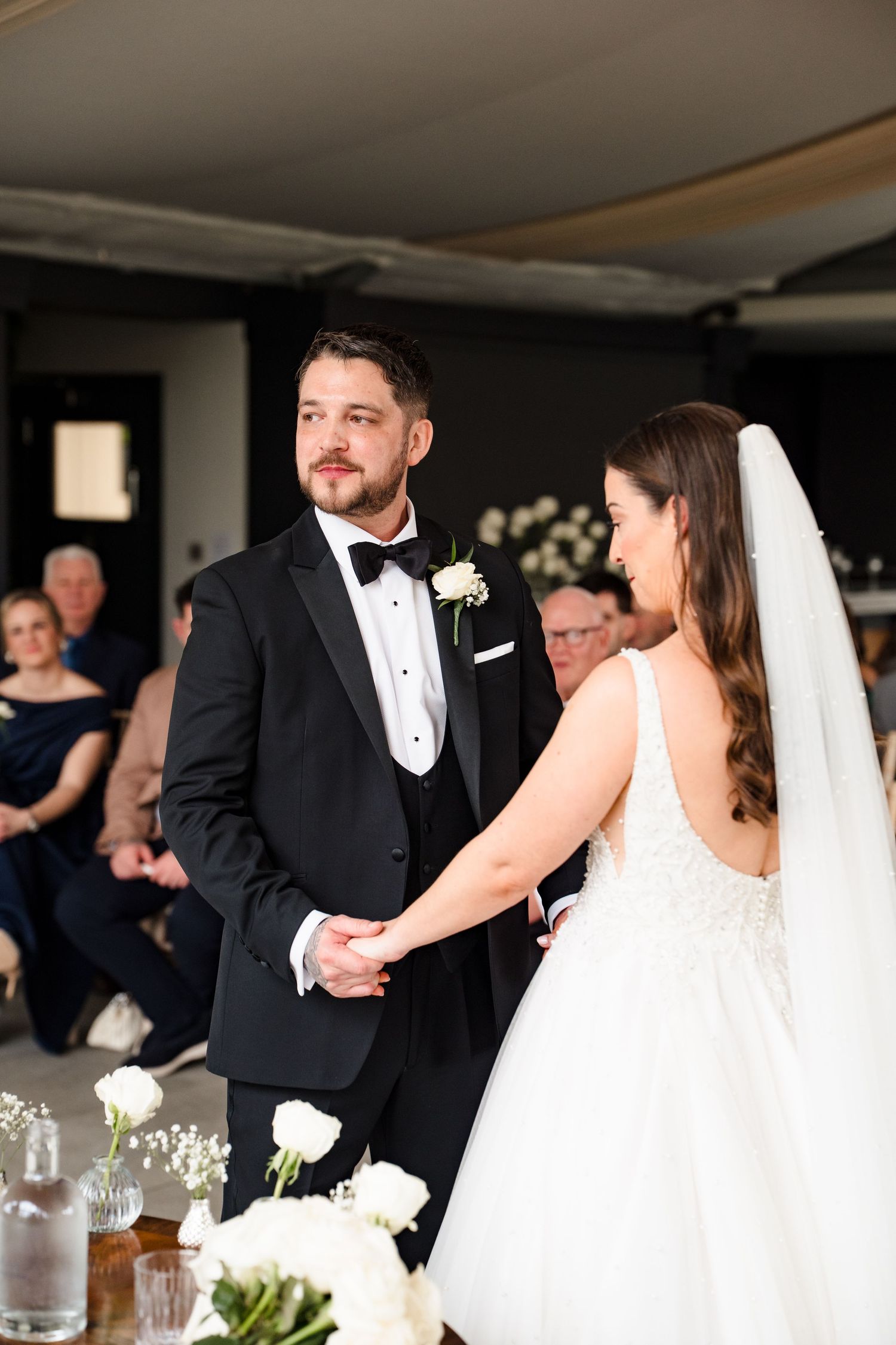 Groom in black tuxedo holds hands with bride in white gown during indoor wedding ceremony.