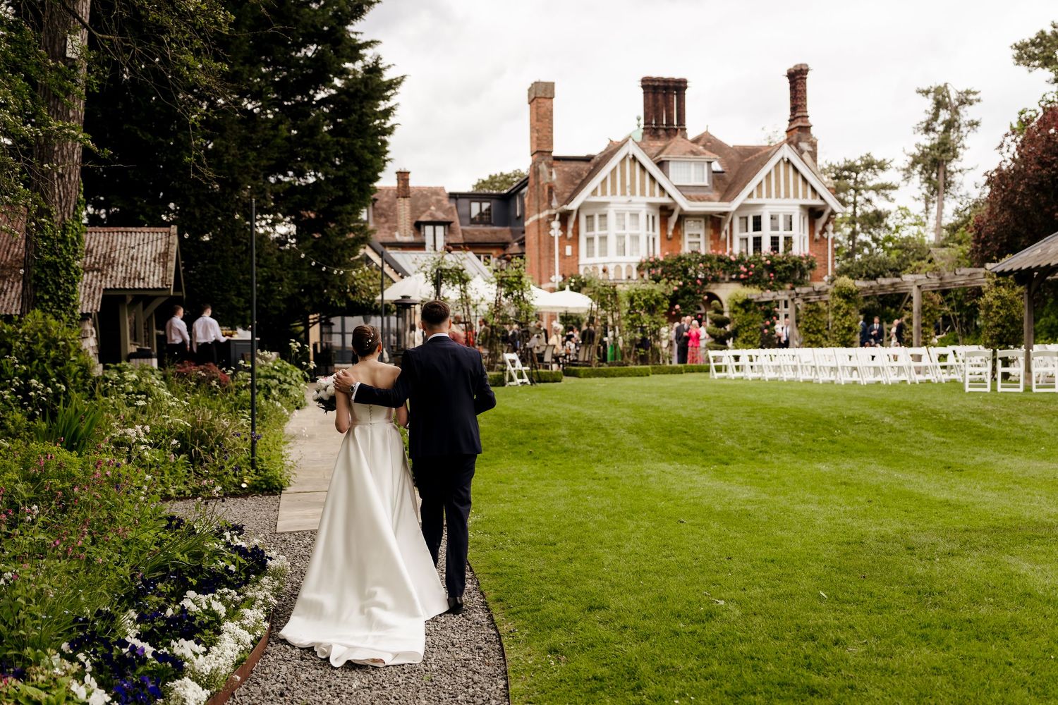 Bride and escort walking towards a grand red-brick wedding venue surrounded by manicured gardens.