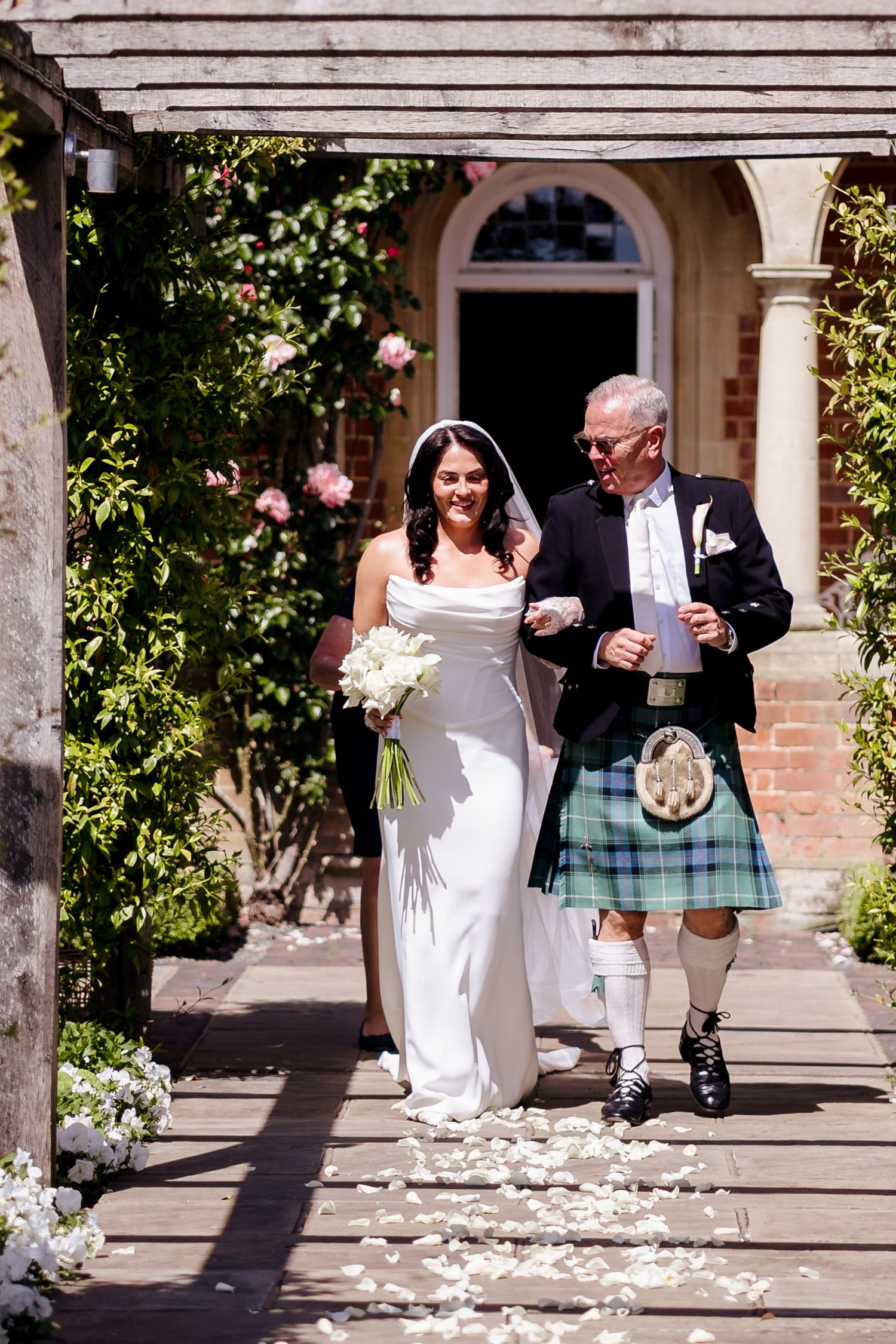 Bride in white gown walks with man in Scottish kilt along petal-strewn aisle at outdoor wedding ceremony.