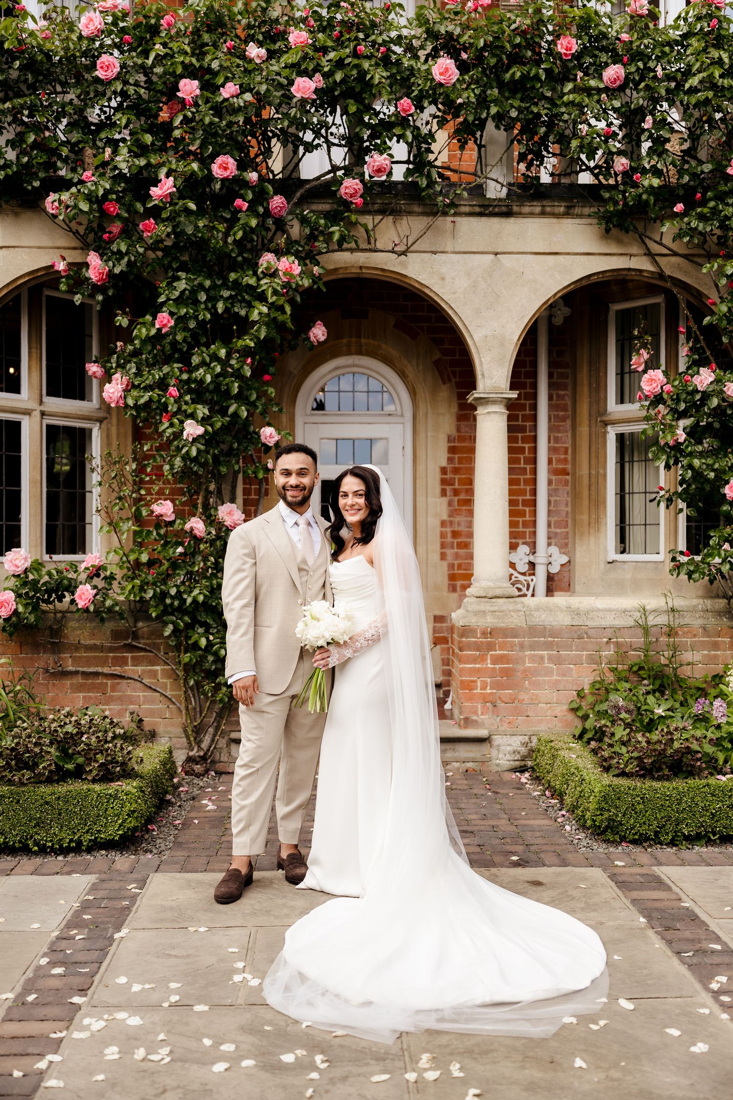 Bride and groom pose before rose-covered brick manor house, bride wearing cathedral veil and white gown.