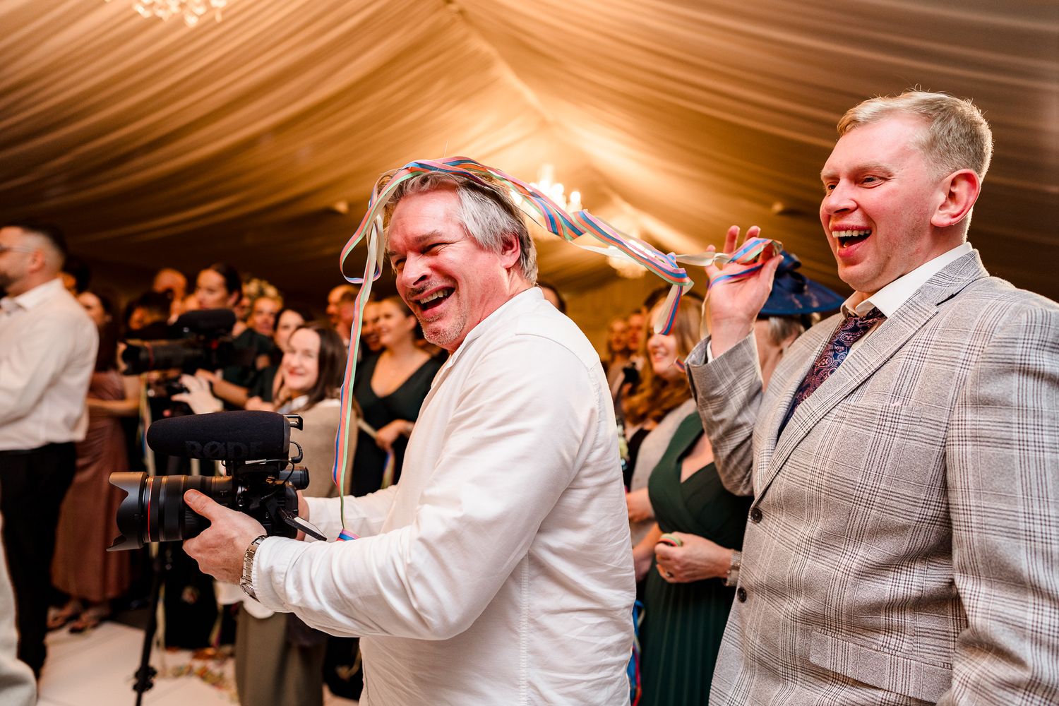 Two men laughing and celebrating joyfully at a wedding reception in a marquee venue.
