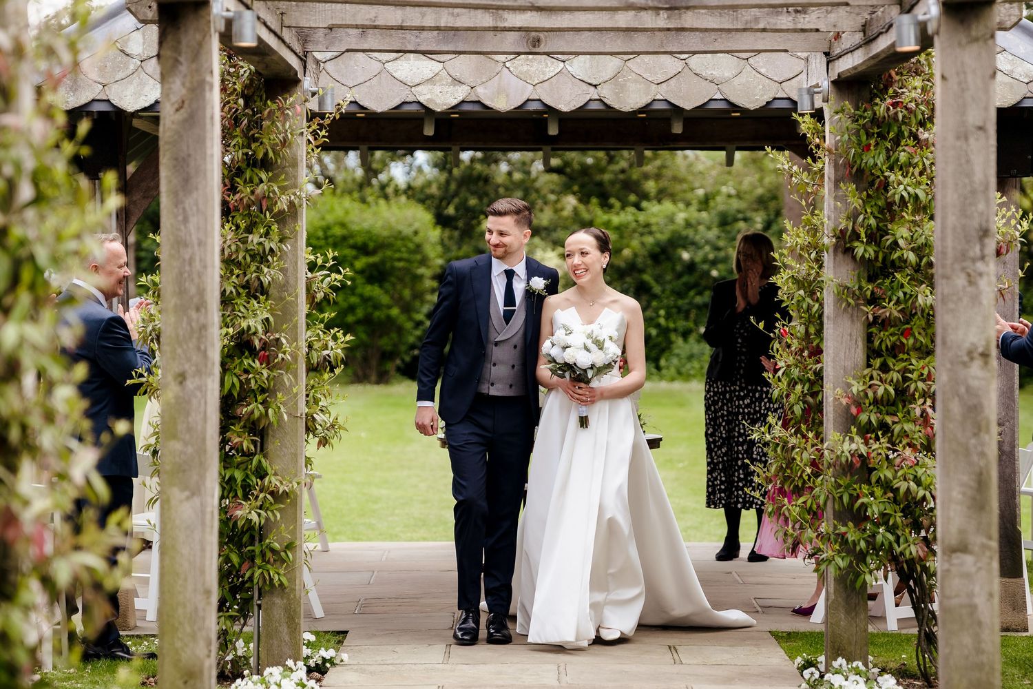 Bride and groom walking through a floral pergola archway on their wedding day in a lush garden setting.
