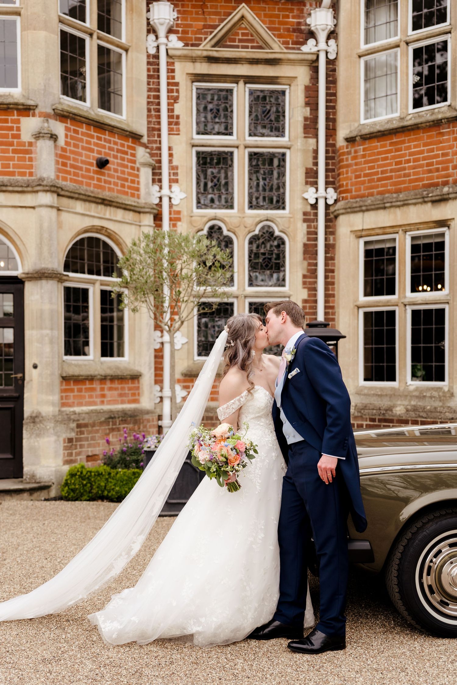 Bride and groom kiss beside a vintage car in front of a grand red-brick wedding venue.