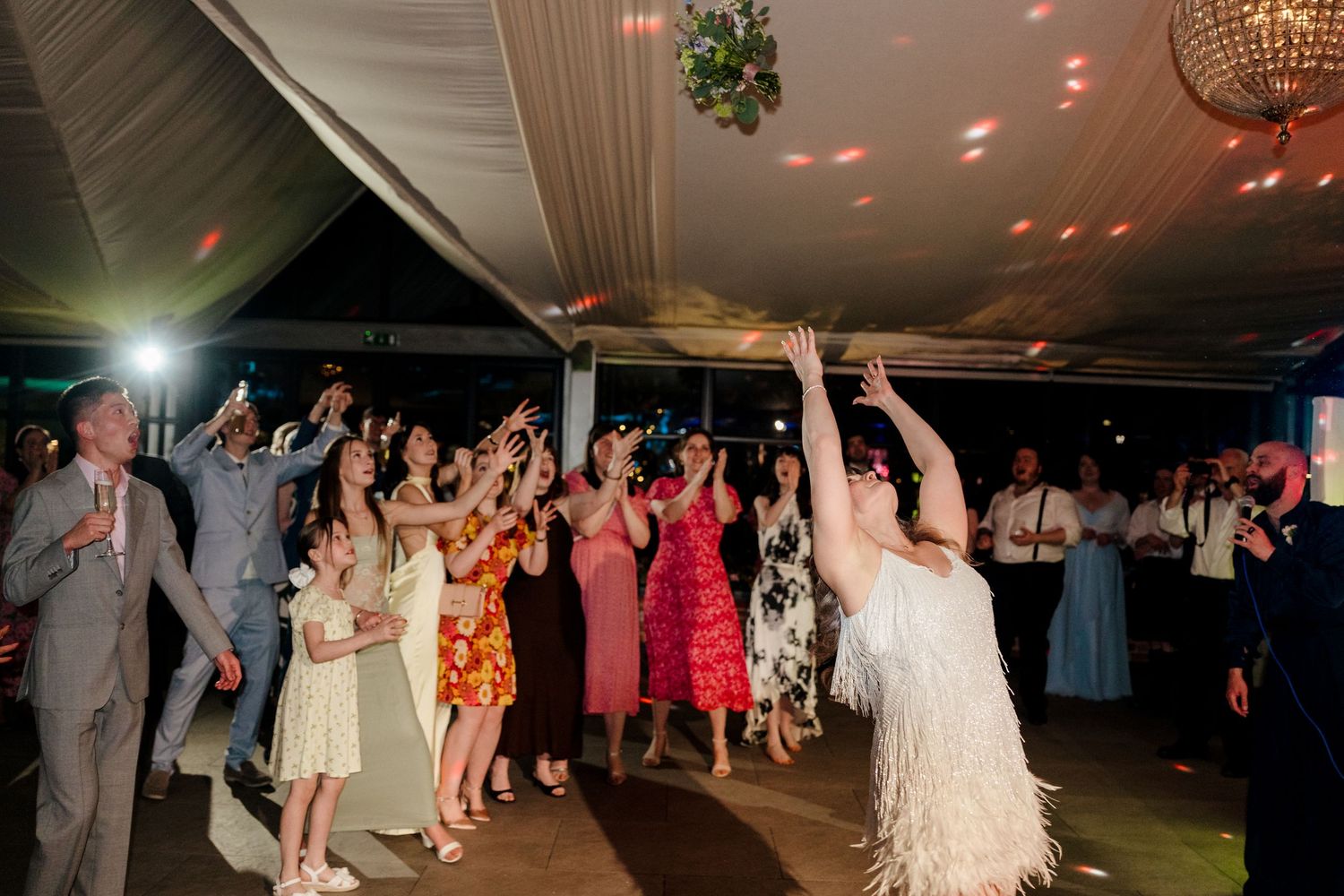 Bride in a feathered dress tosses her bouquet to excited guests on the wedding dance floor.