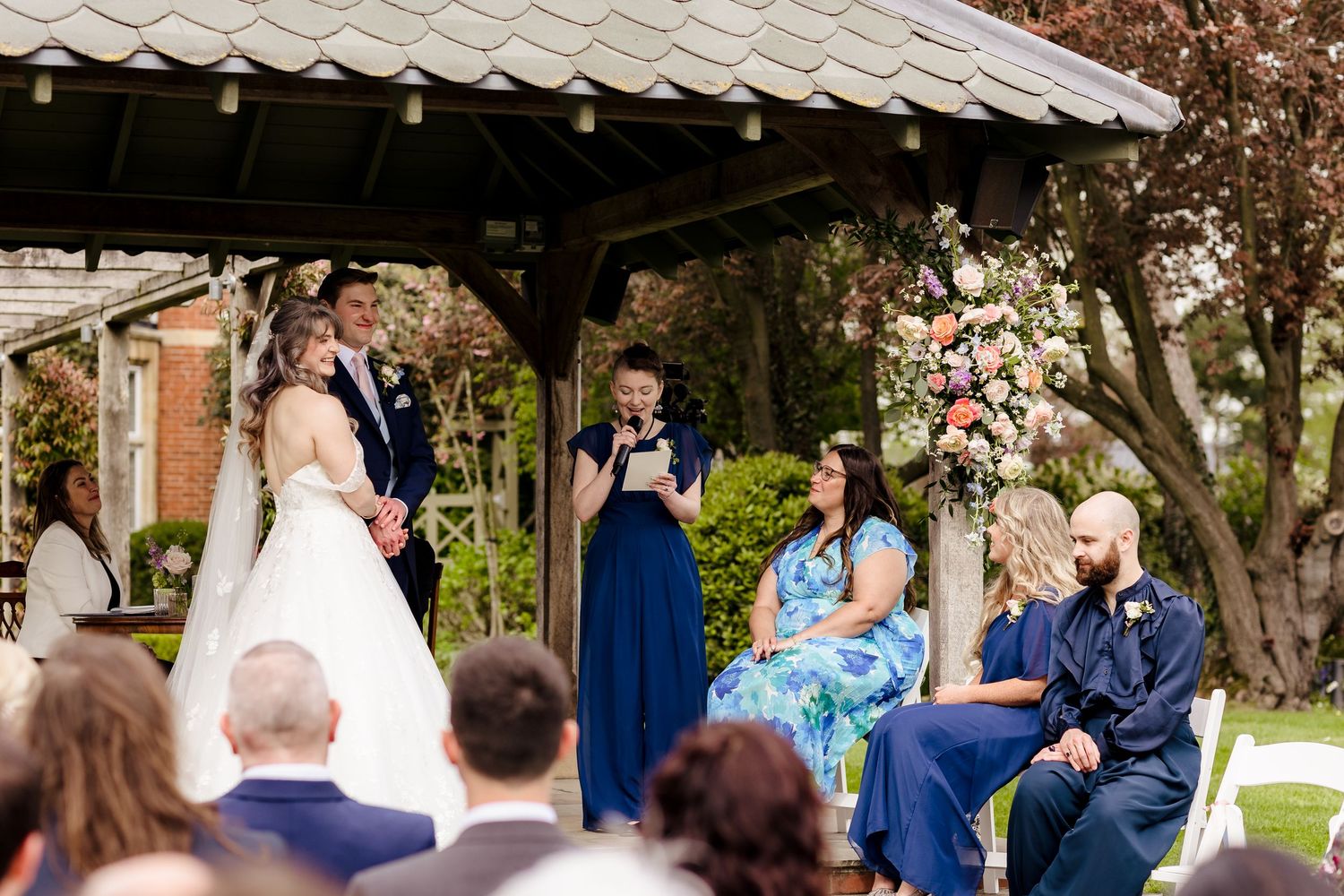 Wedding ceremony under gazebo with bride, groom, and officiant surrounded by floral arrangements.