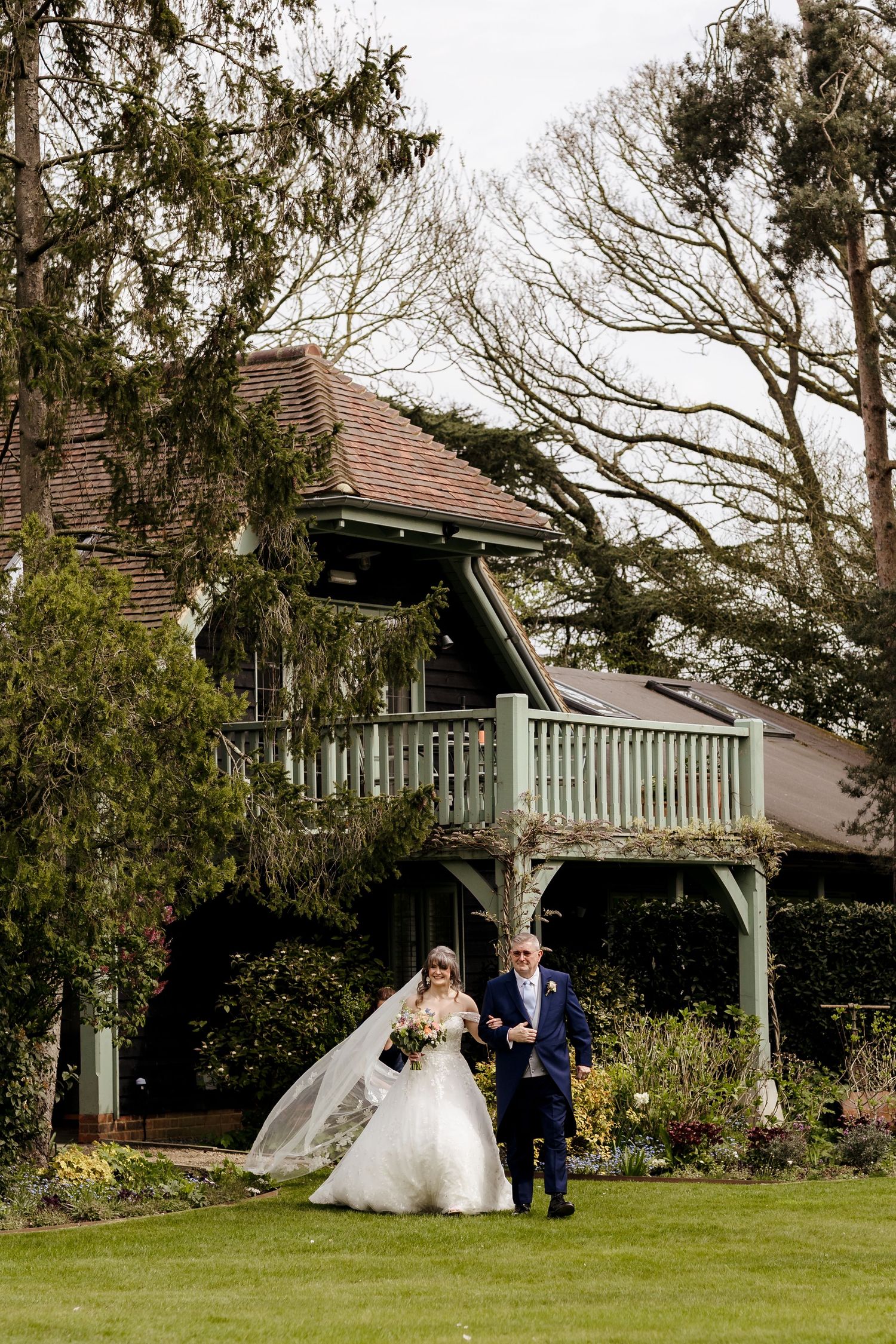 Bride in a flowing white gown walks with escort across garden lawn toward rustic wedding venue.