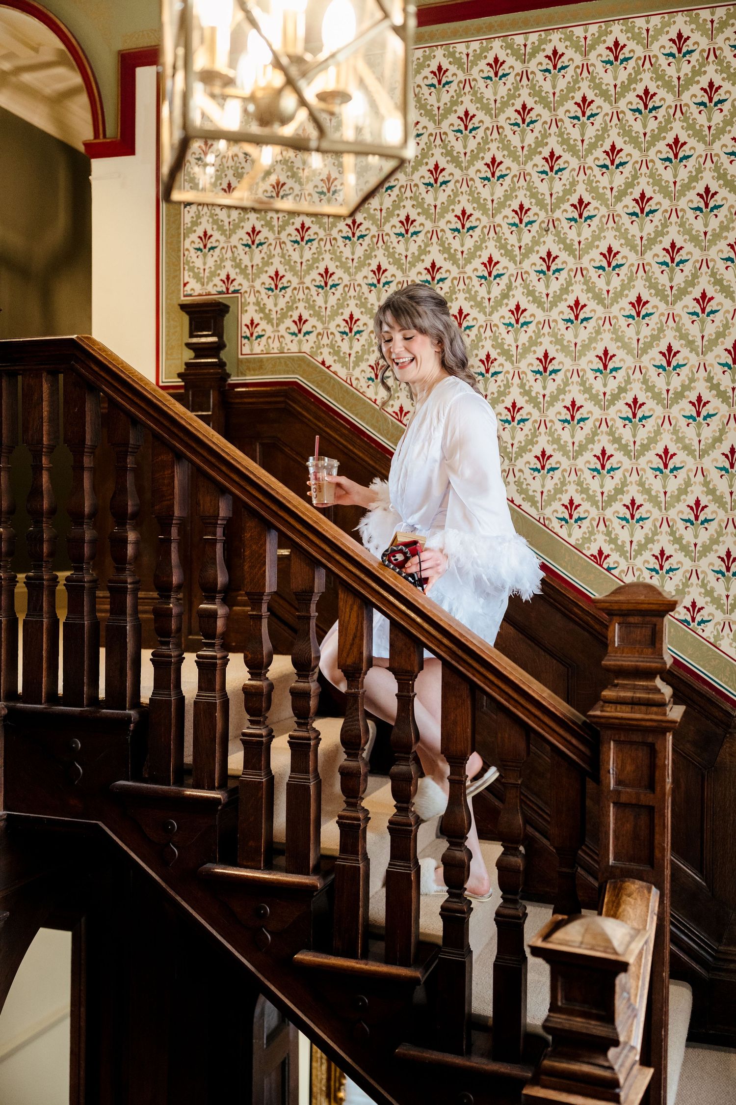 Smiling bride in white robe descending elegant wooden staircase with patterned wallpaper holding a drink.