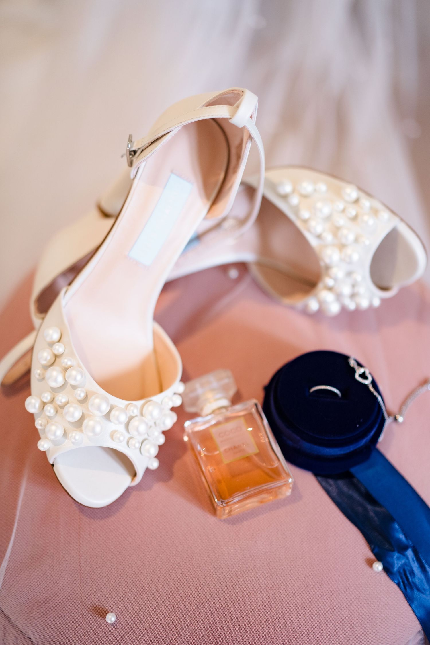 Pearl-embellished bridal heels displayed with perfume bottle, navy garter, and diamond ring on pink surface.