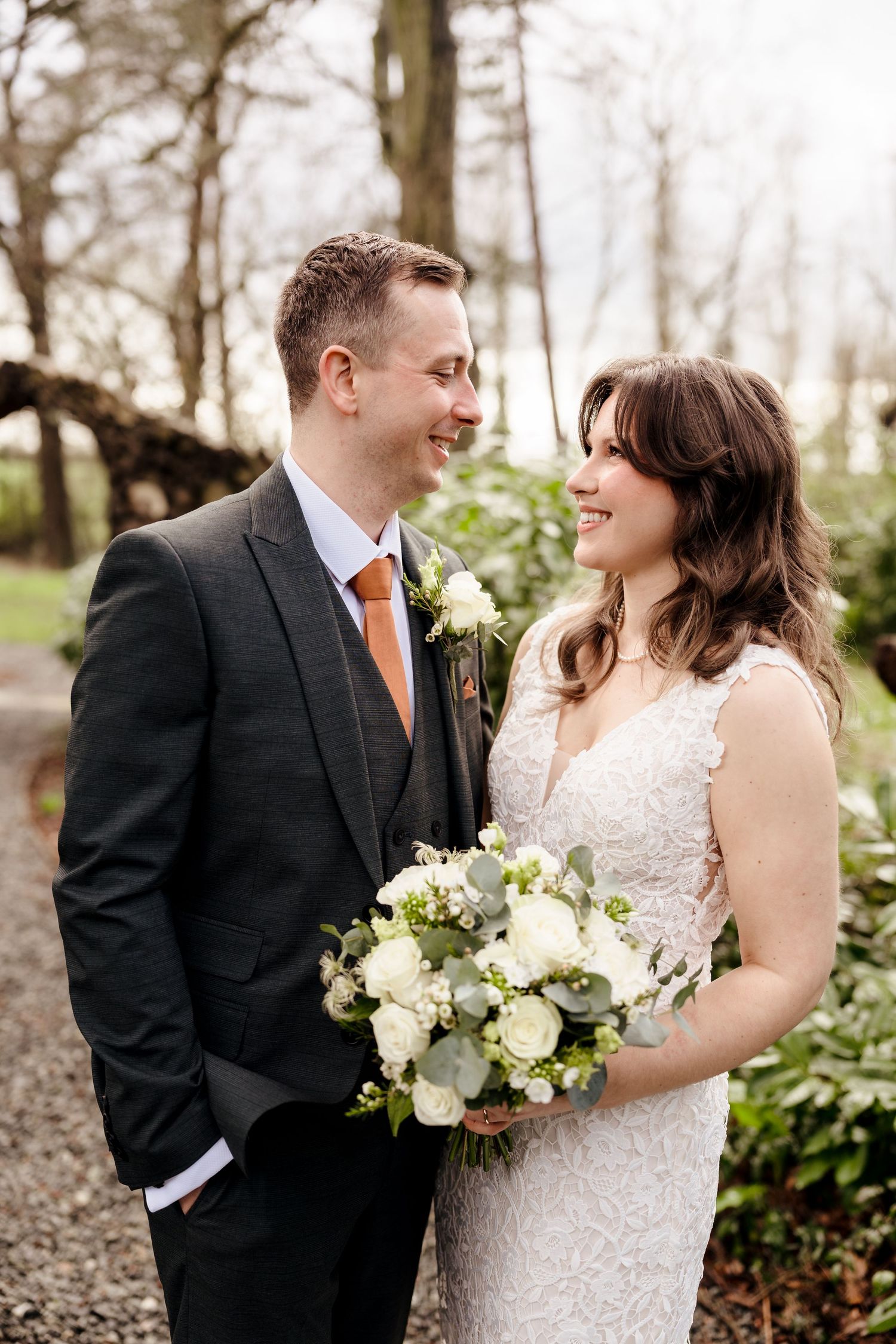 Bride holding white bouquet and groom in grey suit share a smile in a garden setting.