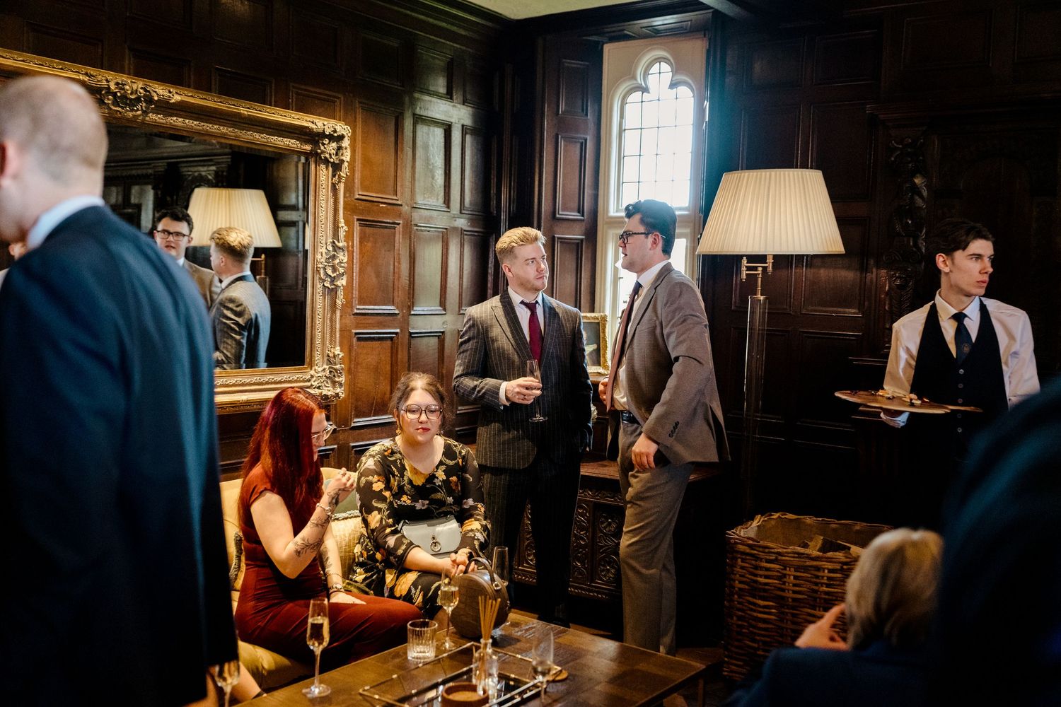 Wedding guests socializing in elegant wood-paneled room with ornate gold mirror.
