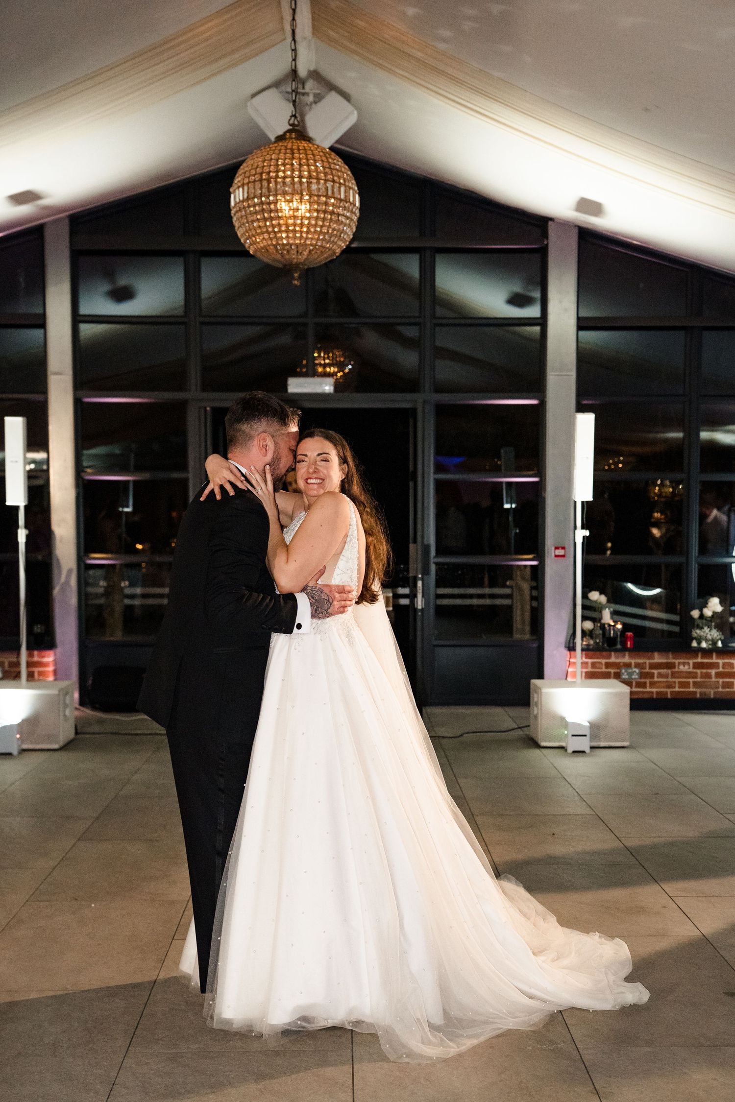 Bride and groom share romantic first dance kiss under a golden chandelier outside venue at night.