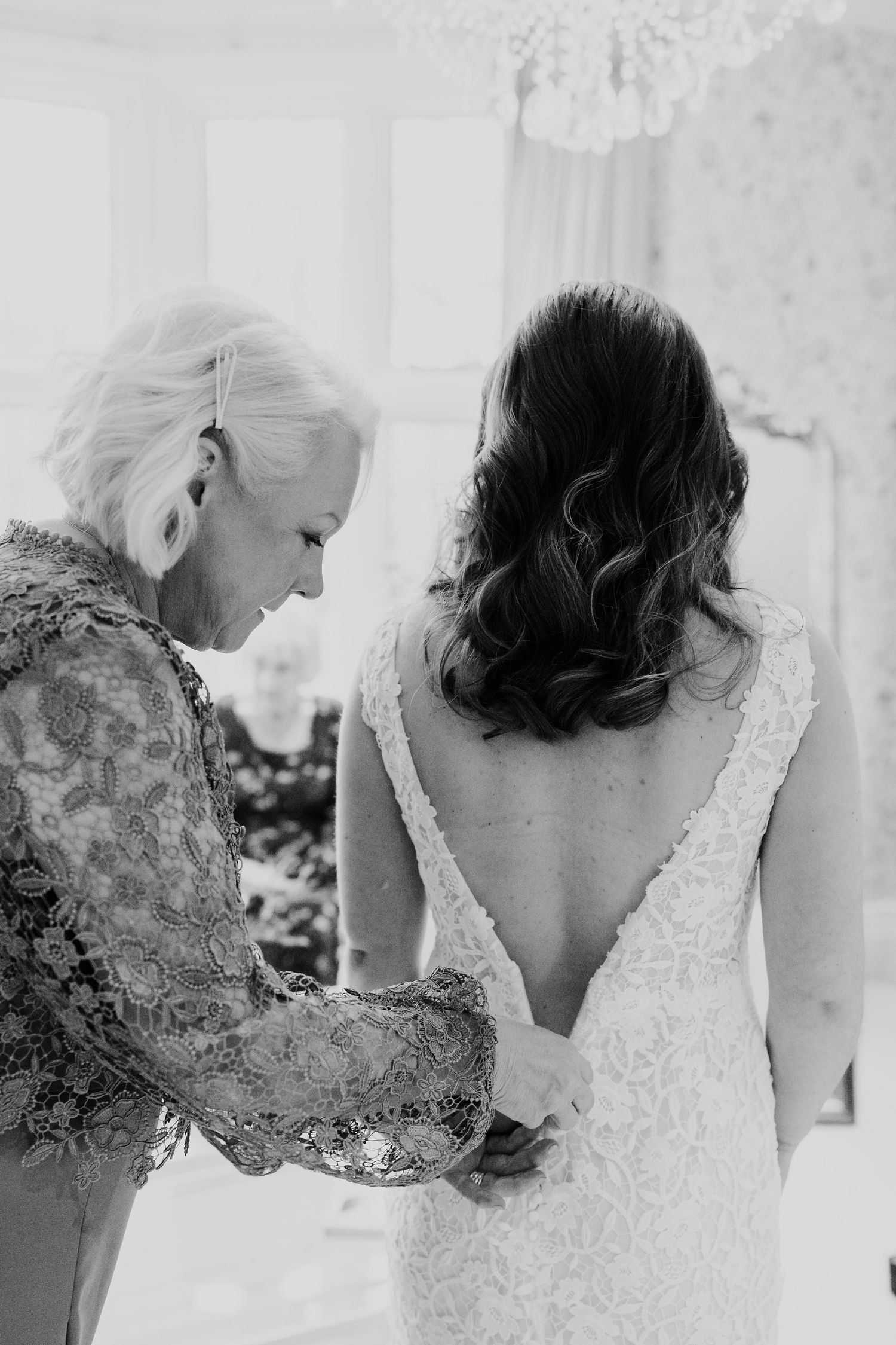 Black and white photo of mother buttoning bride's lace wedding dress with deep V-back detail.