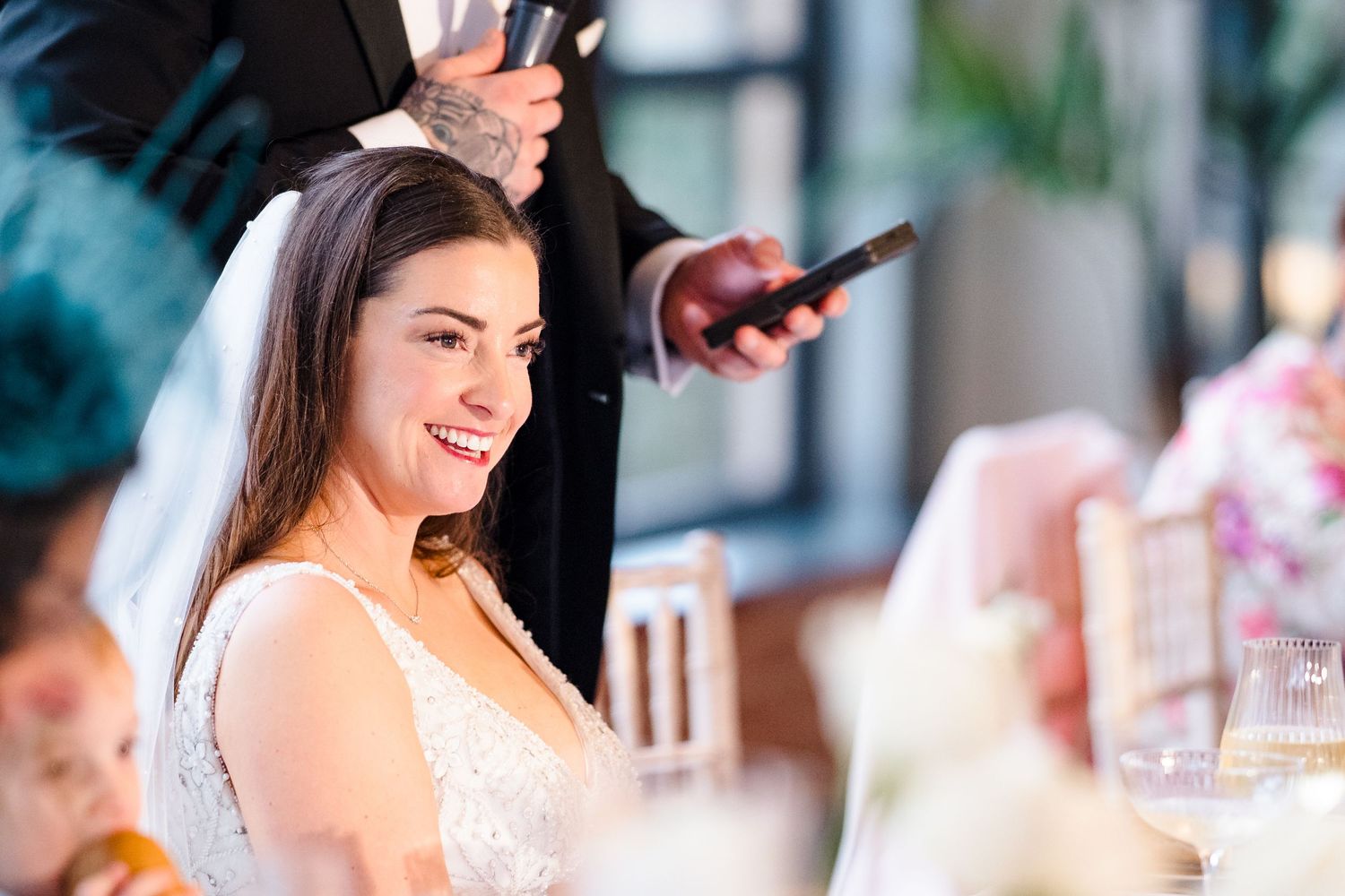 Beautiful bride in lace wedding dress smiling during wedding reception speeches at her table.