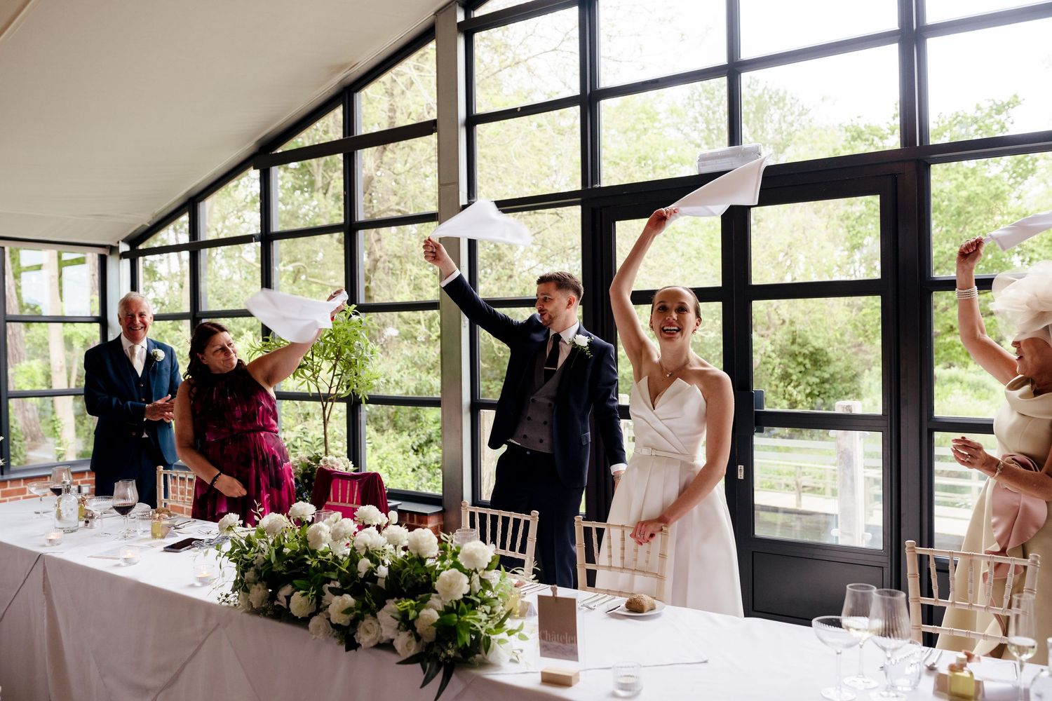 A bride and groom celebrate joyfully waving papers at the wedding reception head table.
