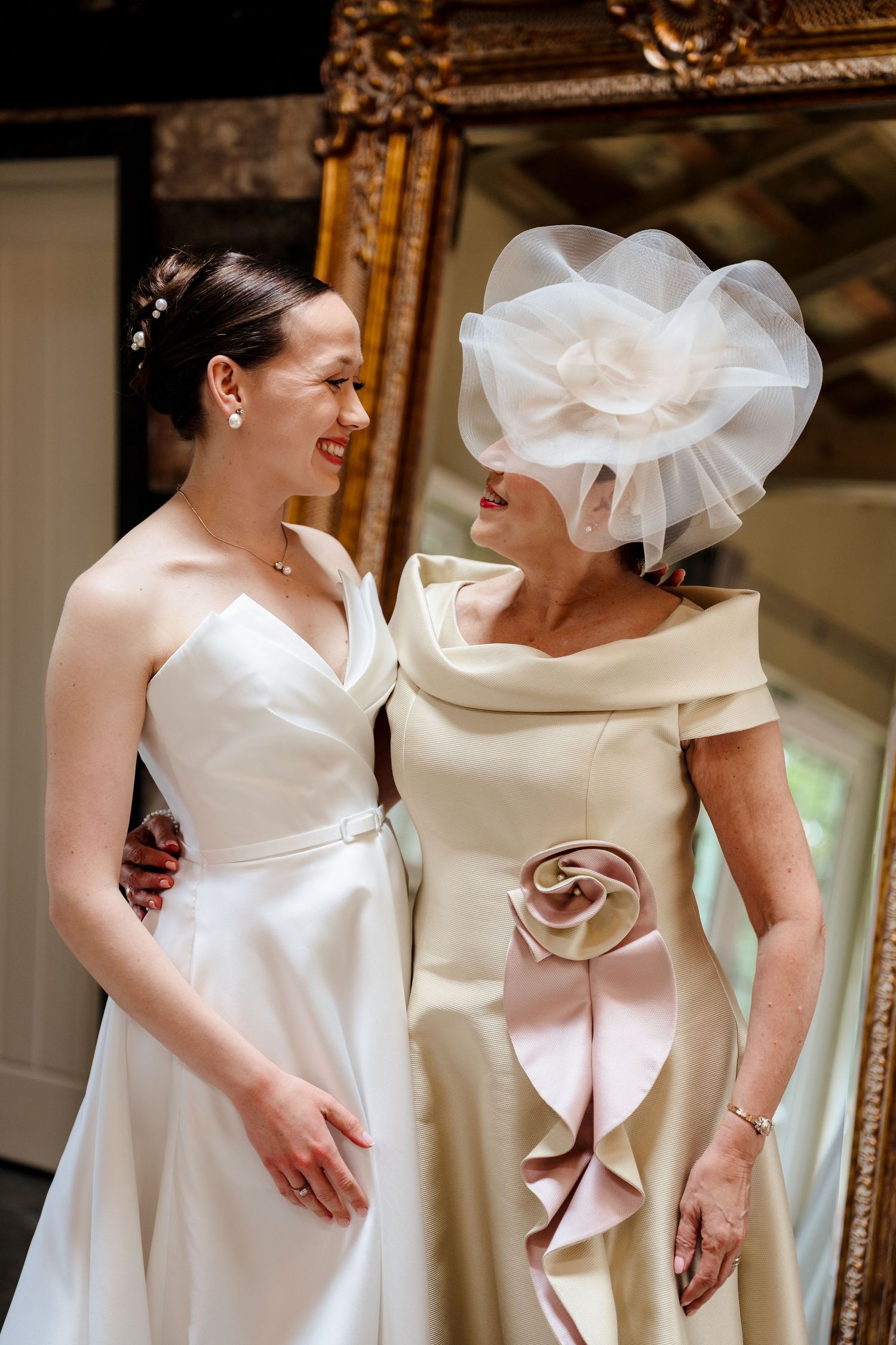 Bride in white strapless gown smiles with older woman in cream dress and white fascinator hat before wedding ceremony.
