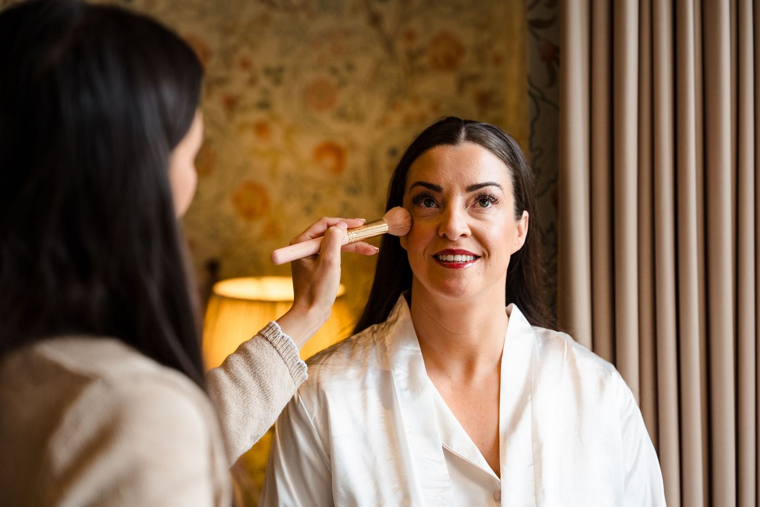 A makeup artist applies foundation with a brush to a smiling woman in a white robe during bridal preparation.